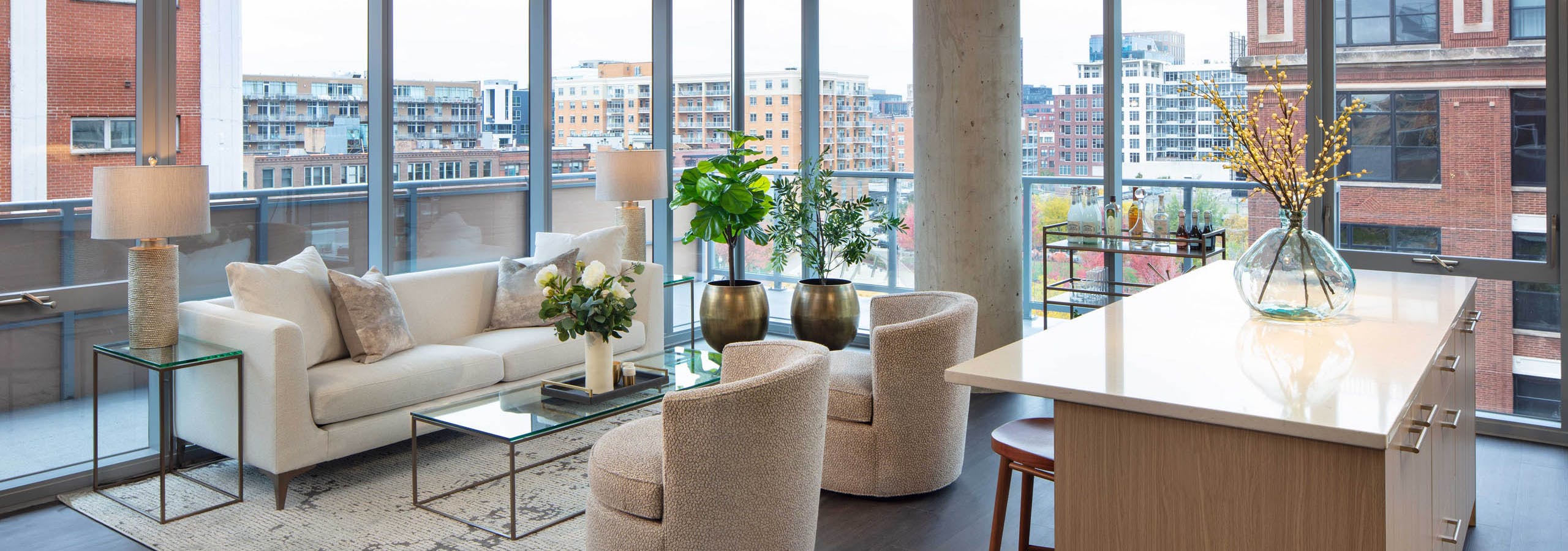 Living room with a white sofa, two textured chairs, a kitchen island, dark flooring, and floor-to-ceiling windows with a city view at AMLI West Loop