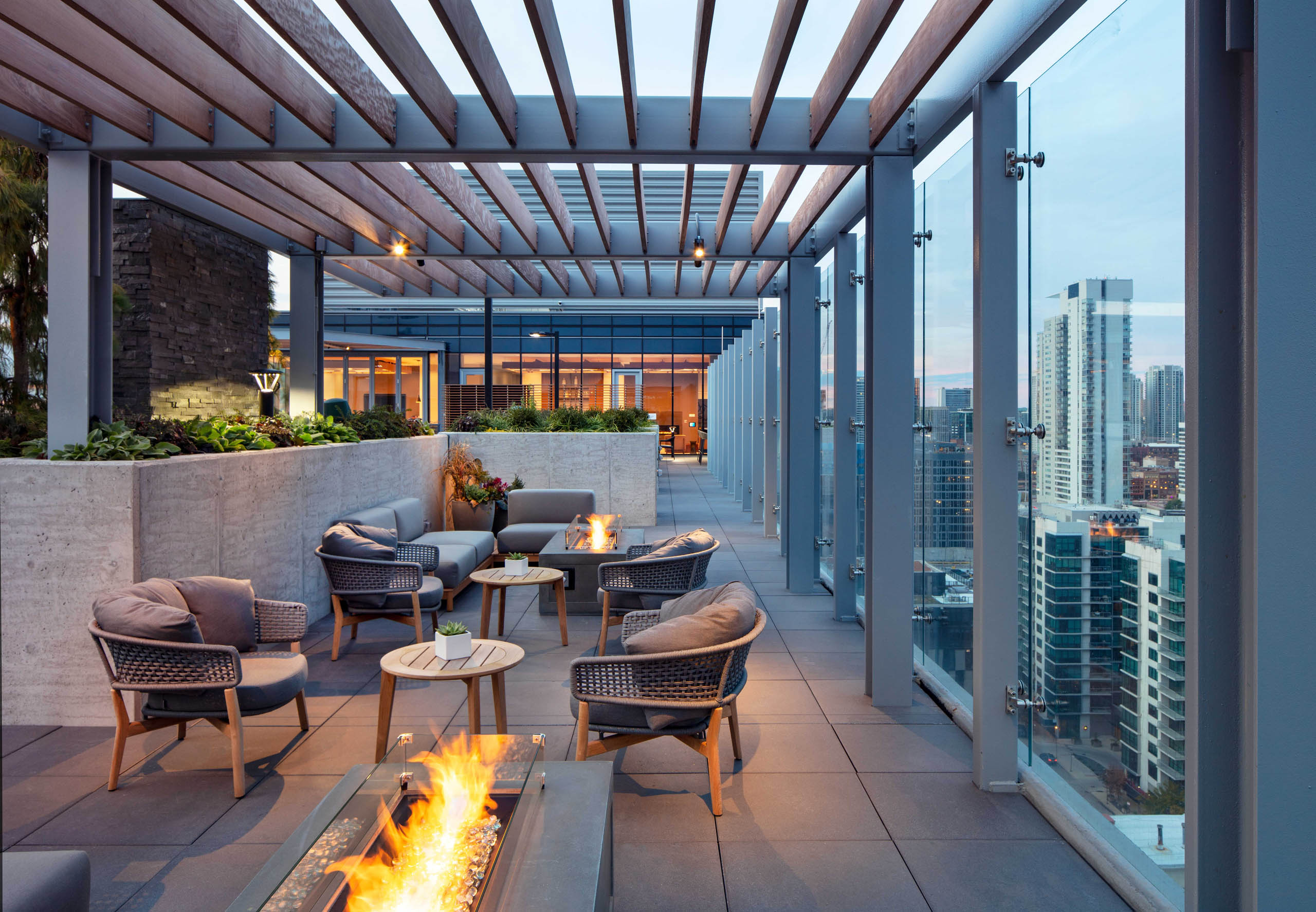 Exterior night shot of AMLI West Loop rooftop terrace with comfortable seating, multiple lit fire pits, and a glass rail overlooking the city skyline.