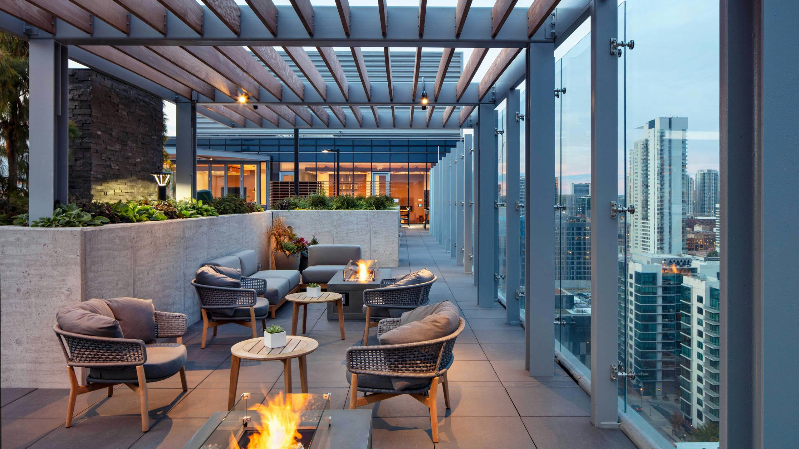 Exterior night shot of AMLI West Loop rooftop terrace with comfortable seating, multiple lit fire pits, and a glass rail overlooking the city skyline.