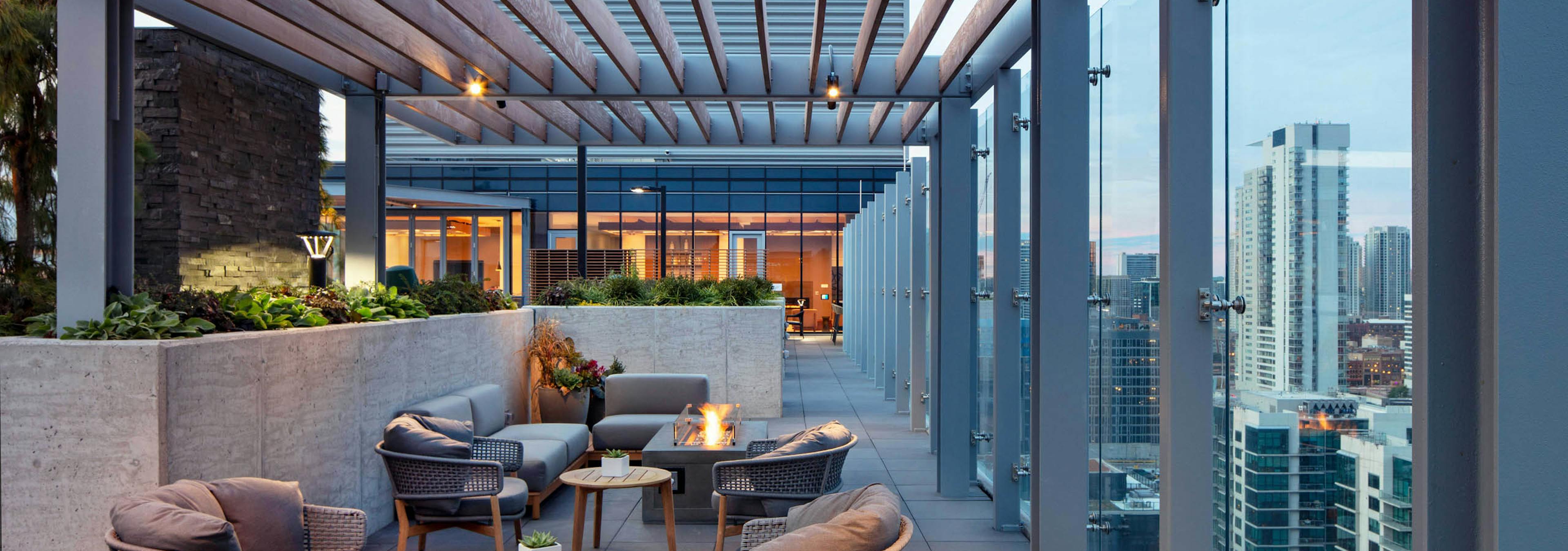 Exterior night shot of AMLI West Loop rooftop terrace with comfortable seating, multiple lit fire pits, and a glass rail overlooking the city skyline.
