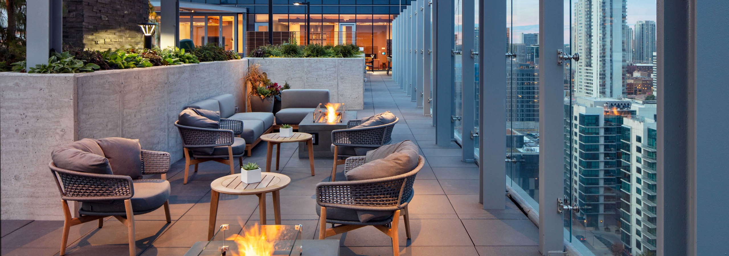 Exterior night shot of AMLI West Loop rooftop terrace with comfortable seating, multiple lit fire pits, and a glass rail overlooking the city skyline.