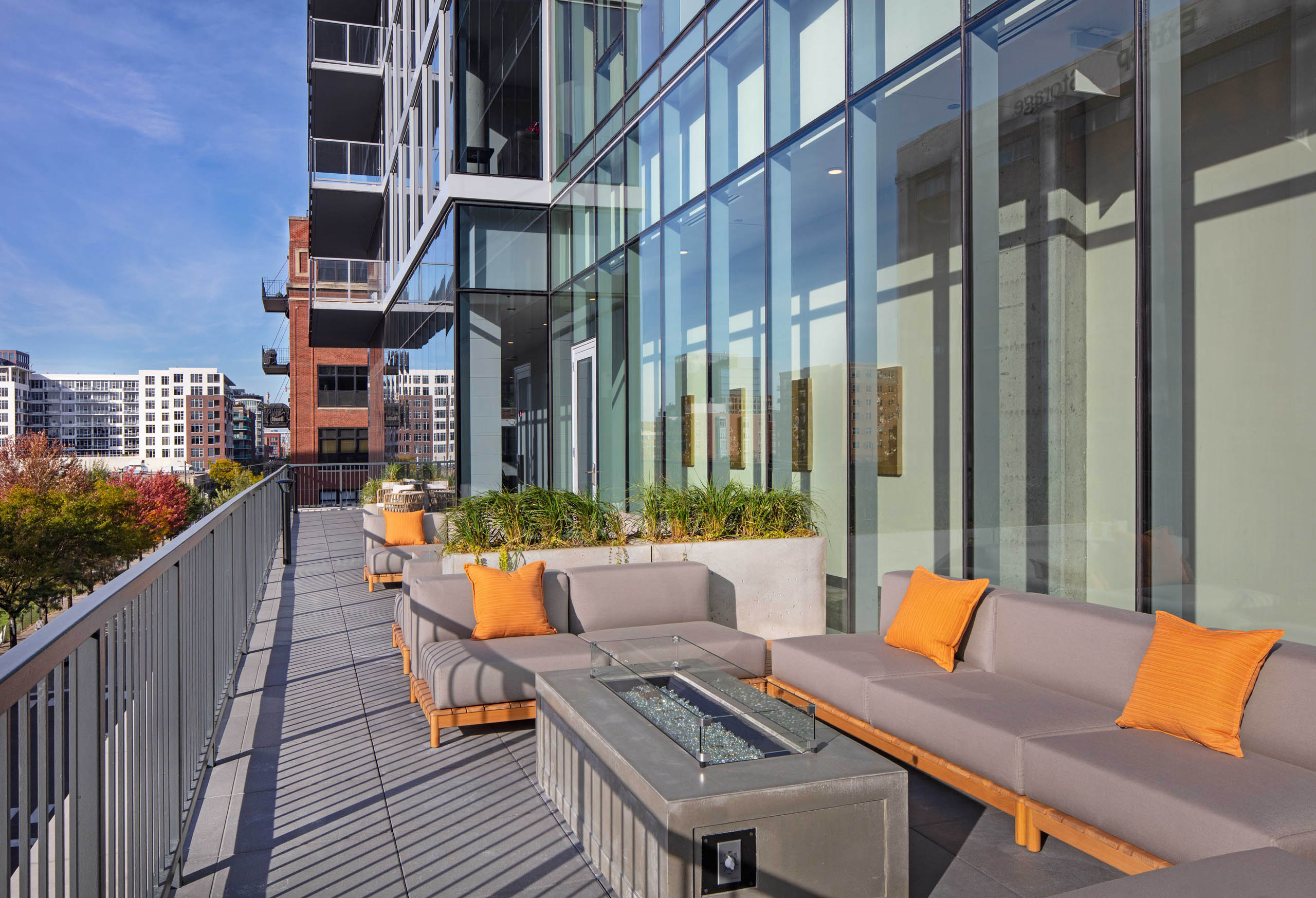 AMLI West Loop firepit deck with modern seating, orange pillows, a fire table, and glass walls overlooking nearby buildings on a sunny day.