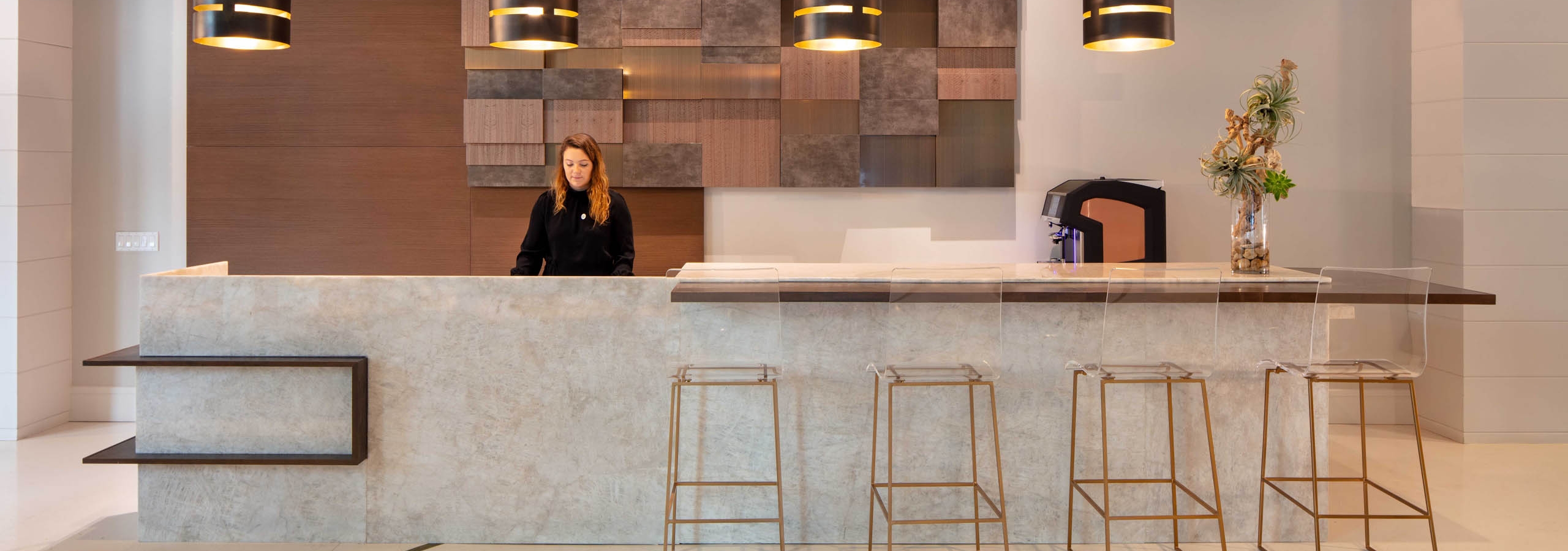 AMLI West Loop modern lobby with a concierge desk, featuring a woman standing behind it, contemporary pendant lights, and decorative wood paneling.