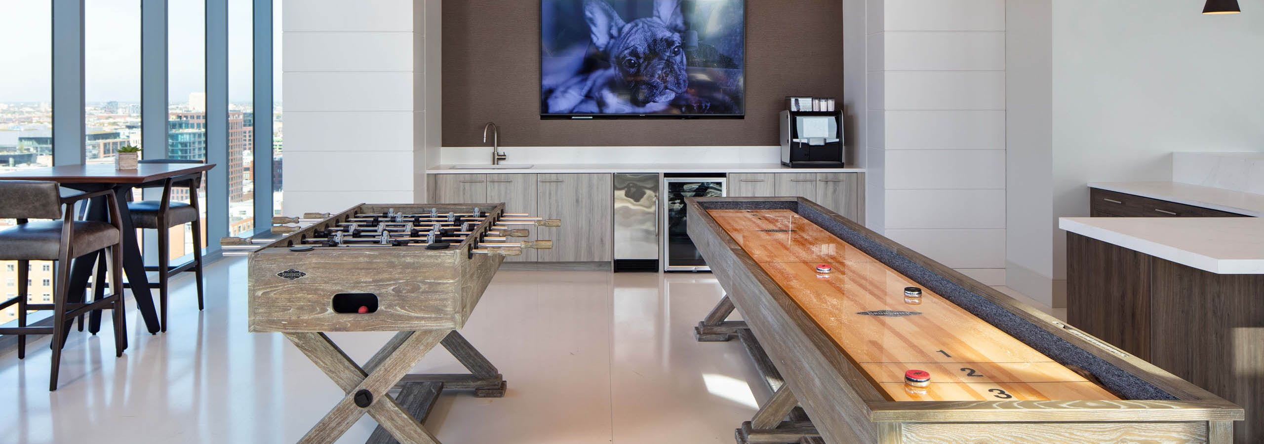 Interior shot of the Resident Lounge game area with foosball and shuffleboard tables, a built-in counter, TV, and large windows at AMLI West Loop