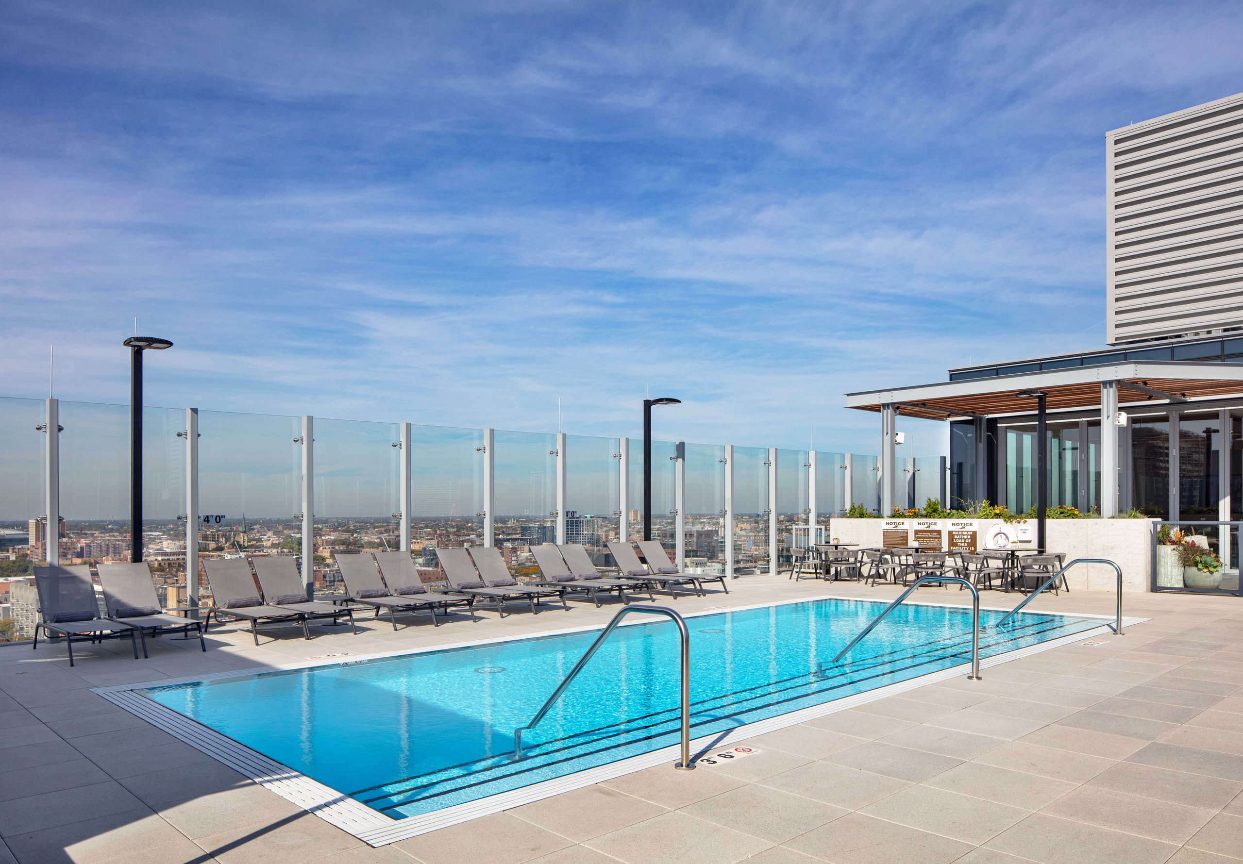 Daytime view of AMLI West Loop rooftop swimming pool with lounge chairs, a glass rail, and a dining area, all overlooking the city skyline.