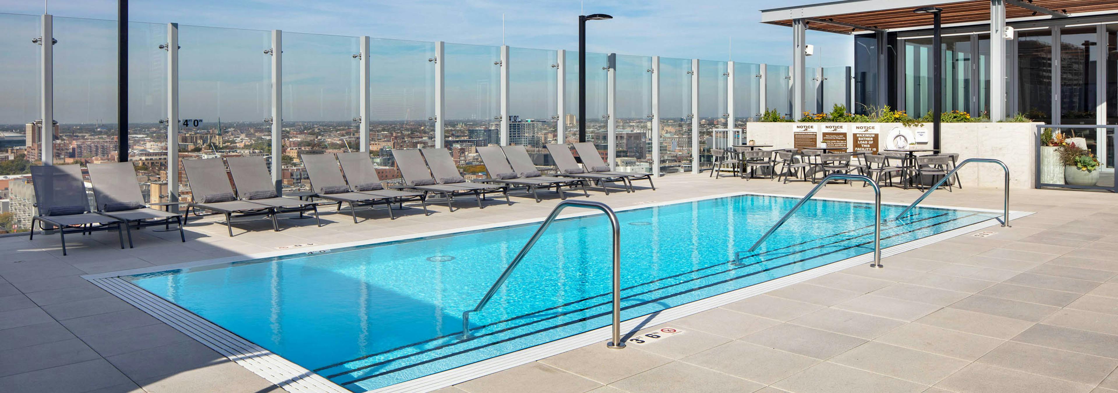 Daytime view of AMLI West Loop rooftop swimming pool with lounge chairs, a glass rail, and a dining area, all overlooking the city skyline.