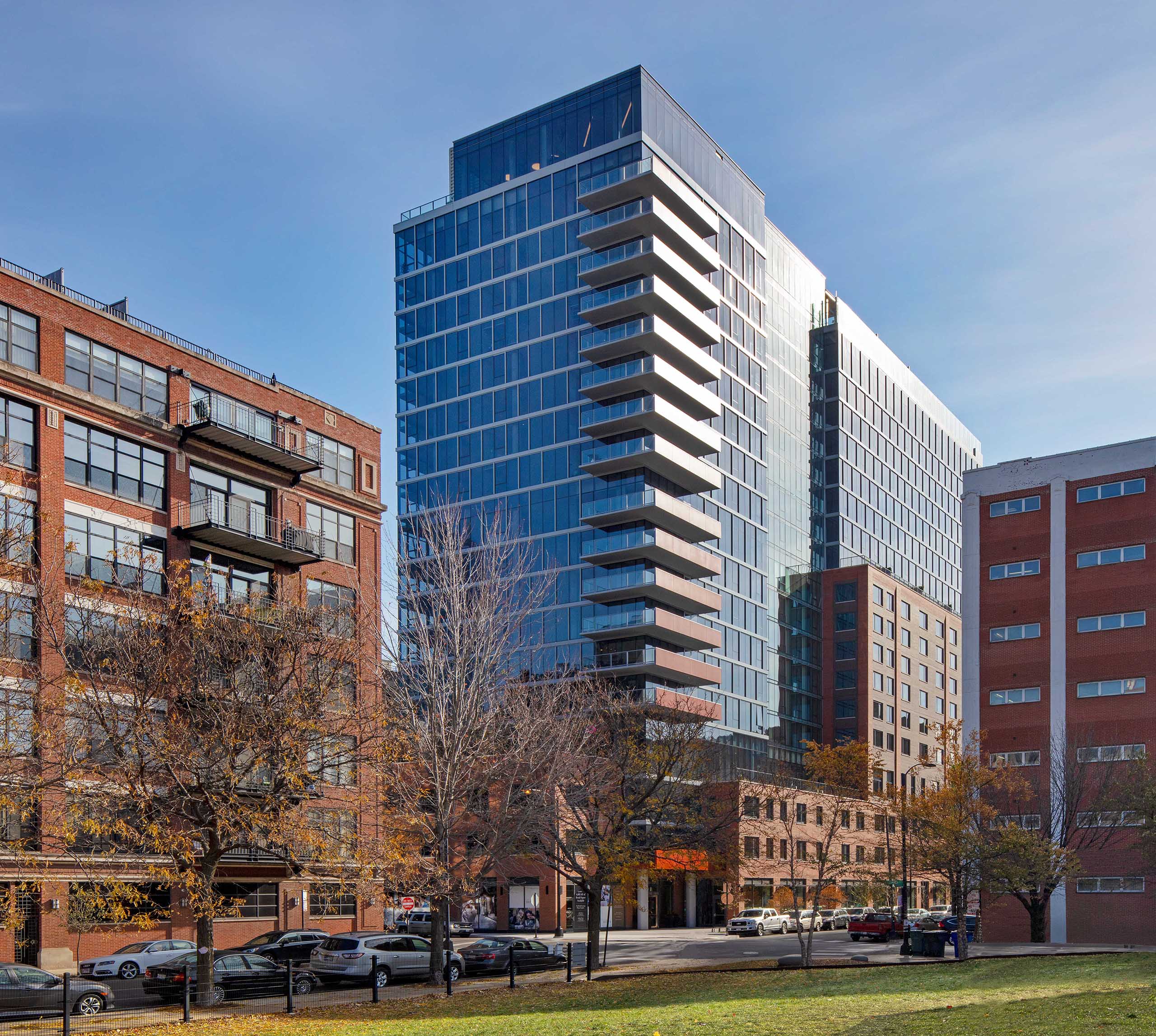 Daytime exterior shot of AMLI West Loop apartment building with surrounding brick buildings on a city street with trees and parked cars.