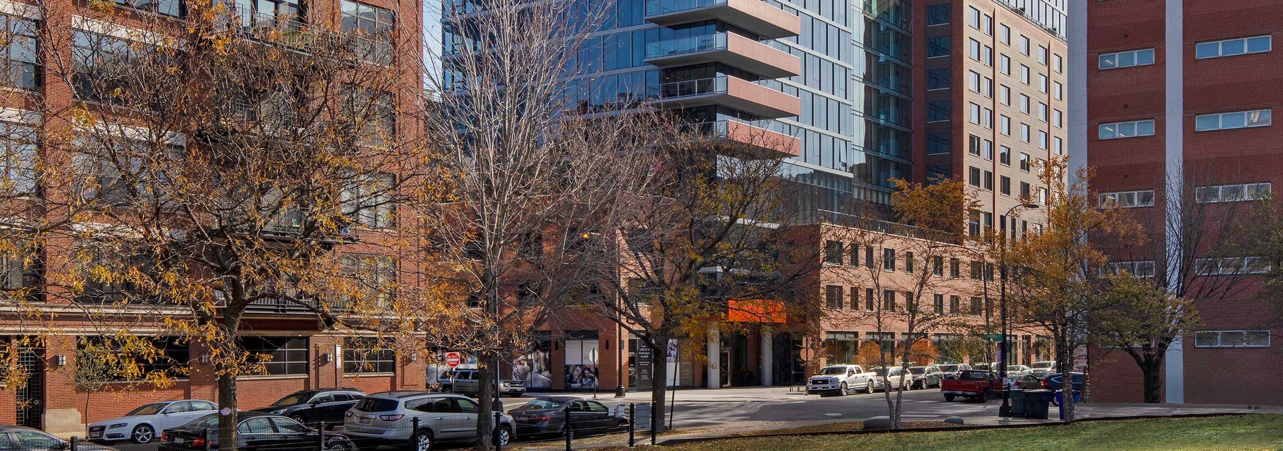 Daytime exterior shot of AMLI West Loop apartment building with surrounding brick buildings on a city street with trees and parked cars.