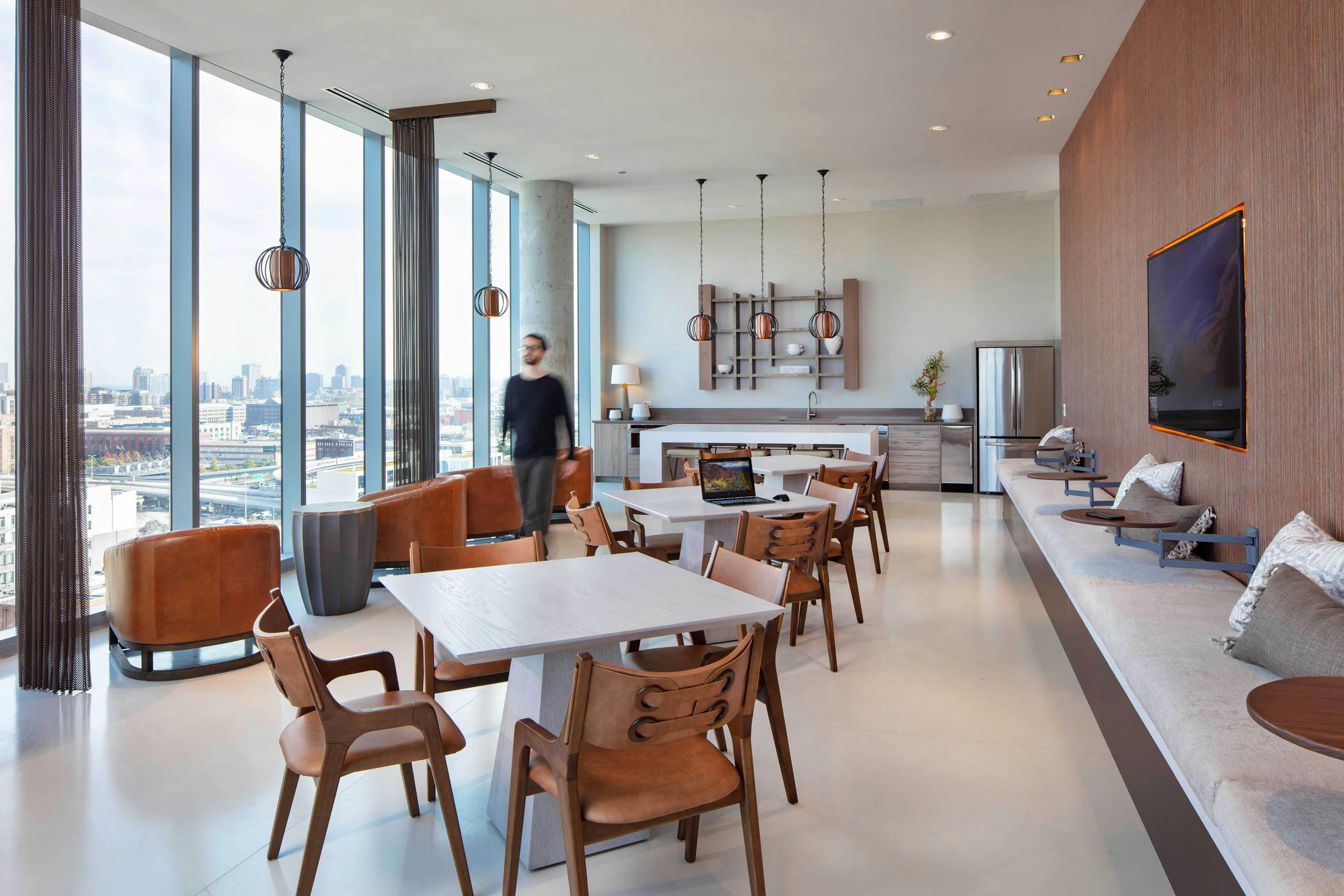 AMLI West Loop social lounge with dining tables, brown leather seating, a kitchen counter, floor-to-ceiling windows, and a man walking past.