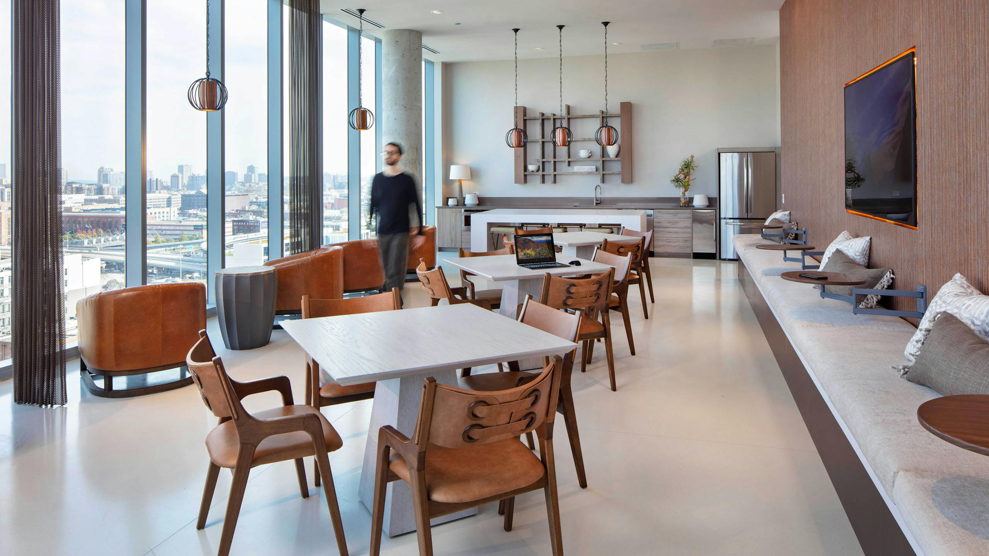 AMLI West Loop social lounge with dining tables, brown leather seating, a kitchen counter, floor-to-ceiling windows, and a man walking past.