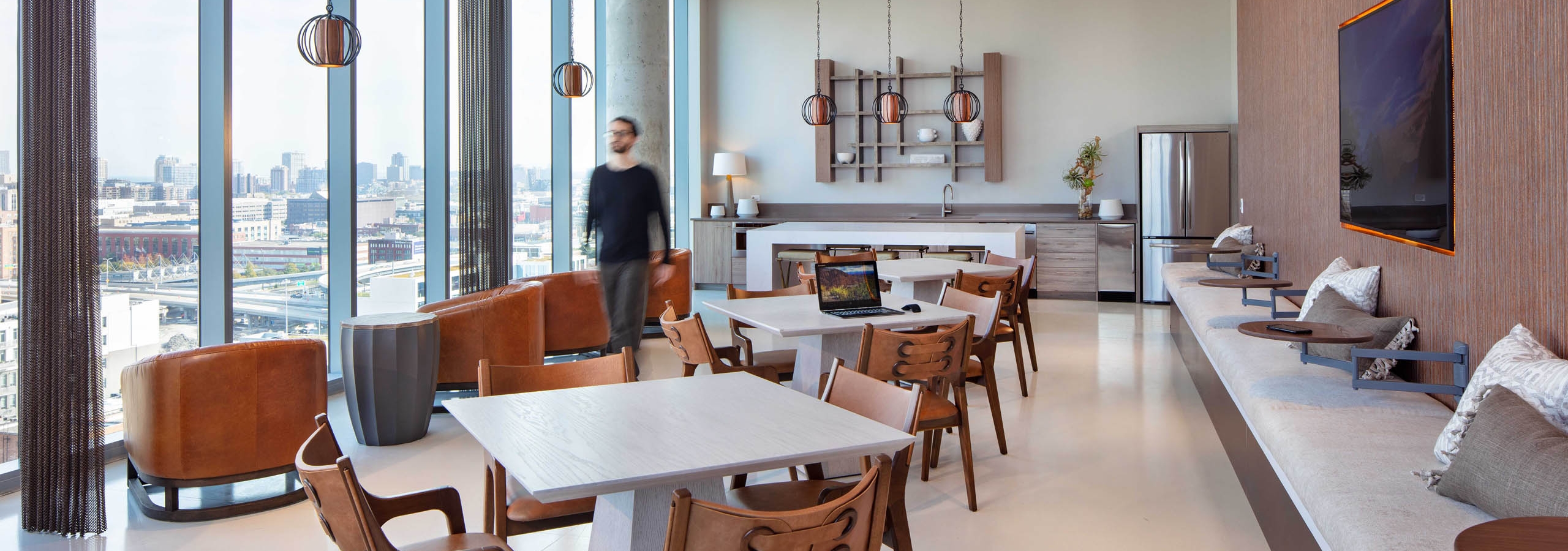 AMLI West Loop social lounge with dining tables, brown leather seating, a kitchen counter, floor-to-ceiling windows, and a man walking past.