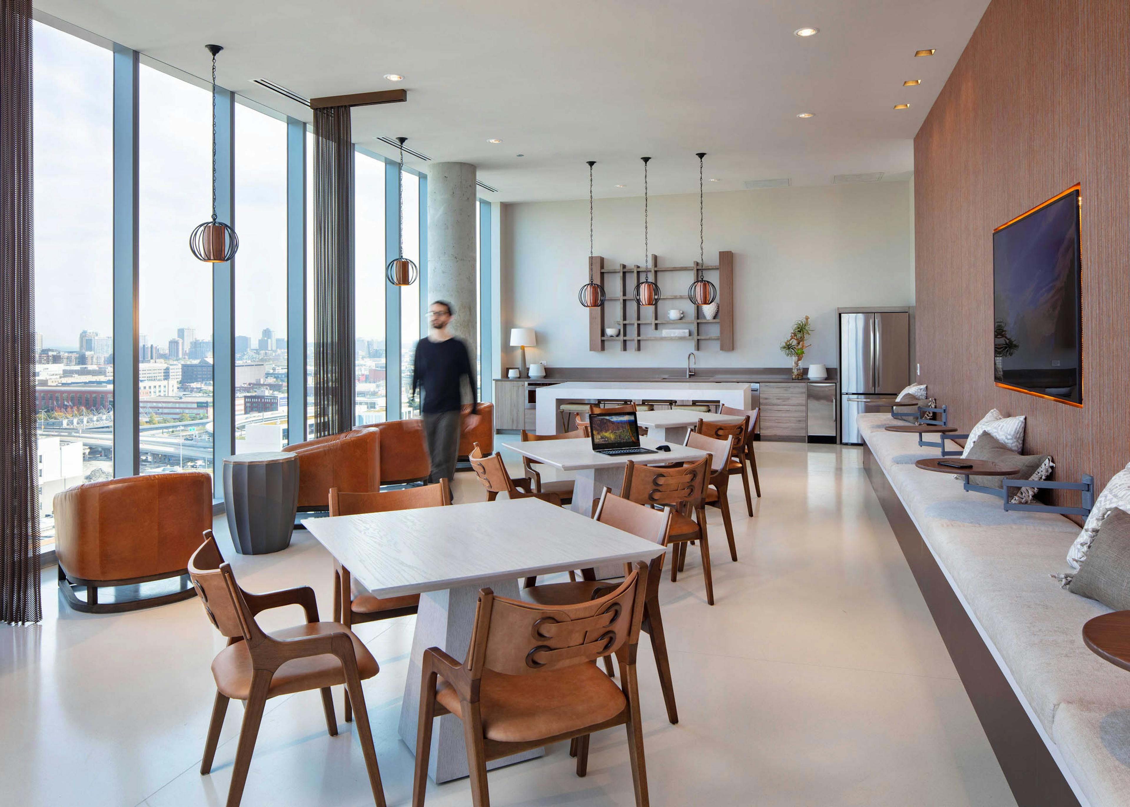 AMLI West Loop social lounge with dining tables, brown leather seating, a kitchen counter, floor-to-ceiling windows, and a man walking past.