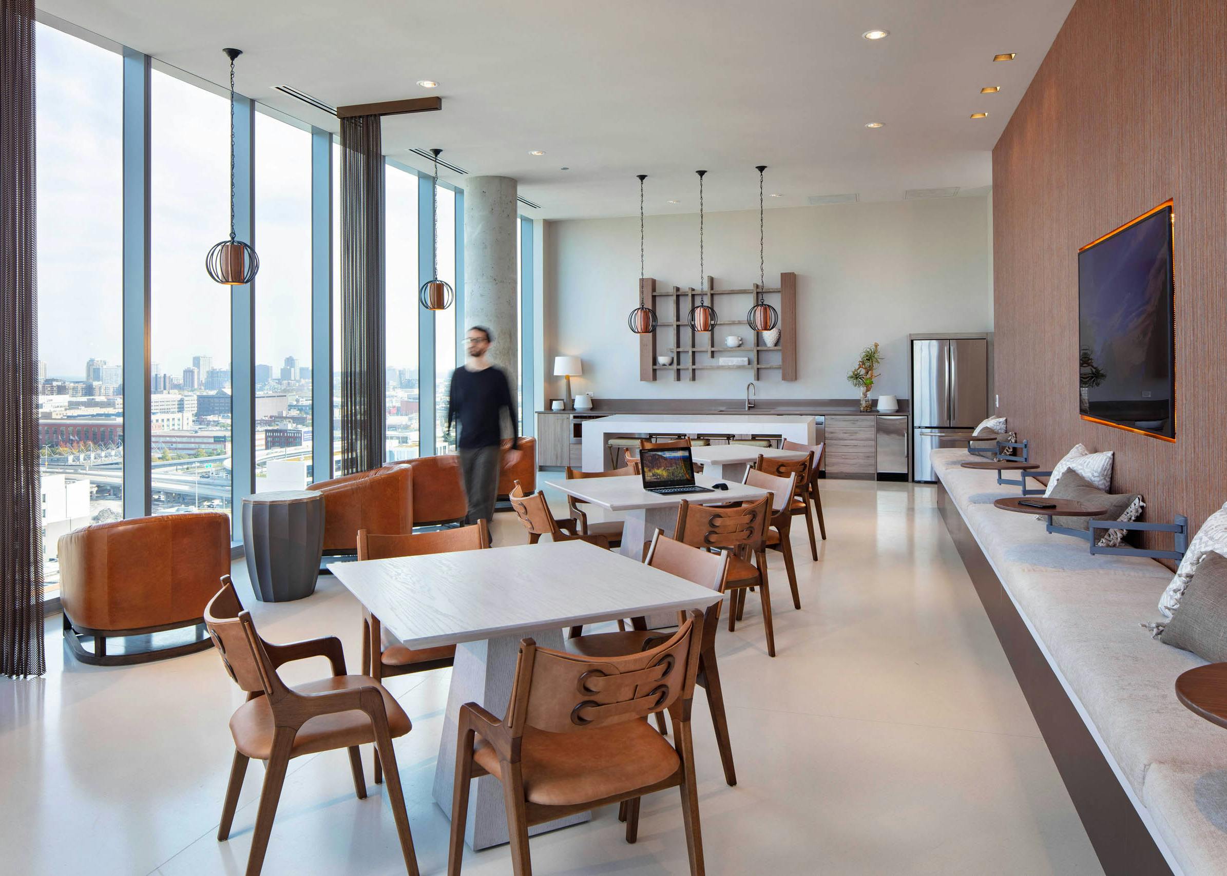 AMLI West Loop social lounge with dining tables, brown leather seating, a kitchen counter, floor-to-ceiling windows, and a man walking past.