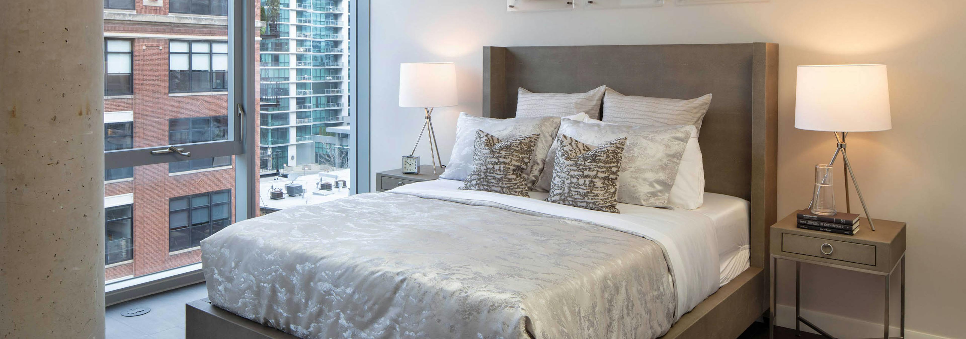 AMLI West Loop bedroom with a large bed, two nightstands and lamps, dark flooring, and floor-to-ceiling windows with a city view.