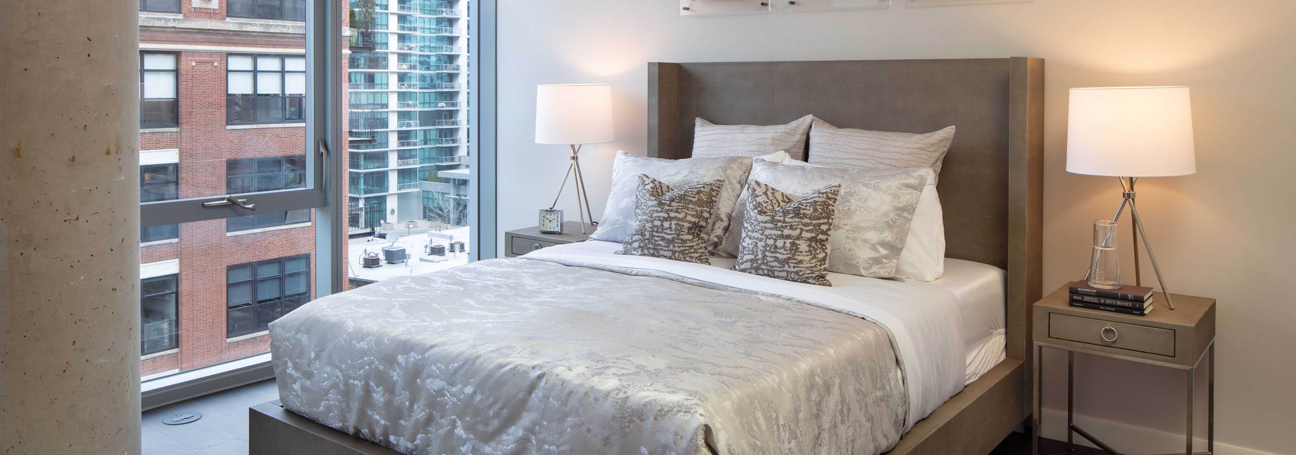 AMLI West Loop bedroom with a large bed, two nightstands and lamps, dark flooring, and floor-to-ceiling windows with a city view.