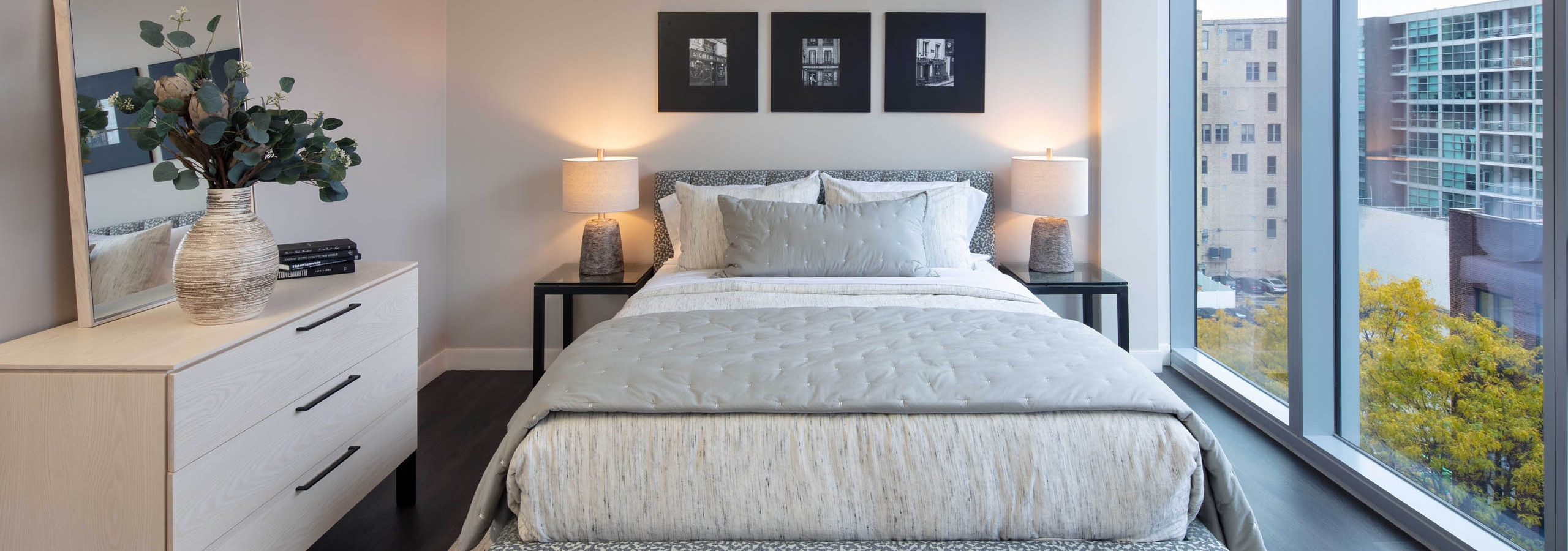 Interior shot of AMLI West Loop bedroom with a queen-sized bed, white dresser with mirror, dark wood flooring, and large window with a city view.