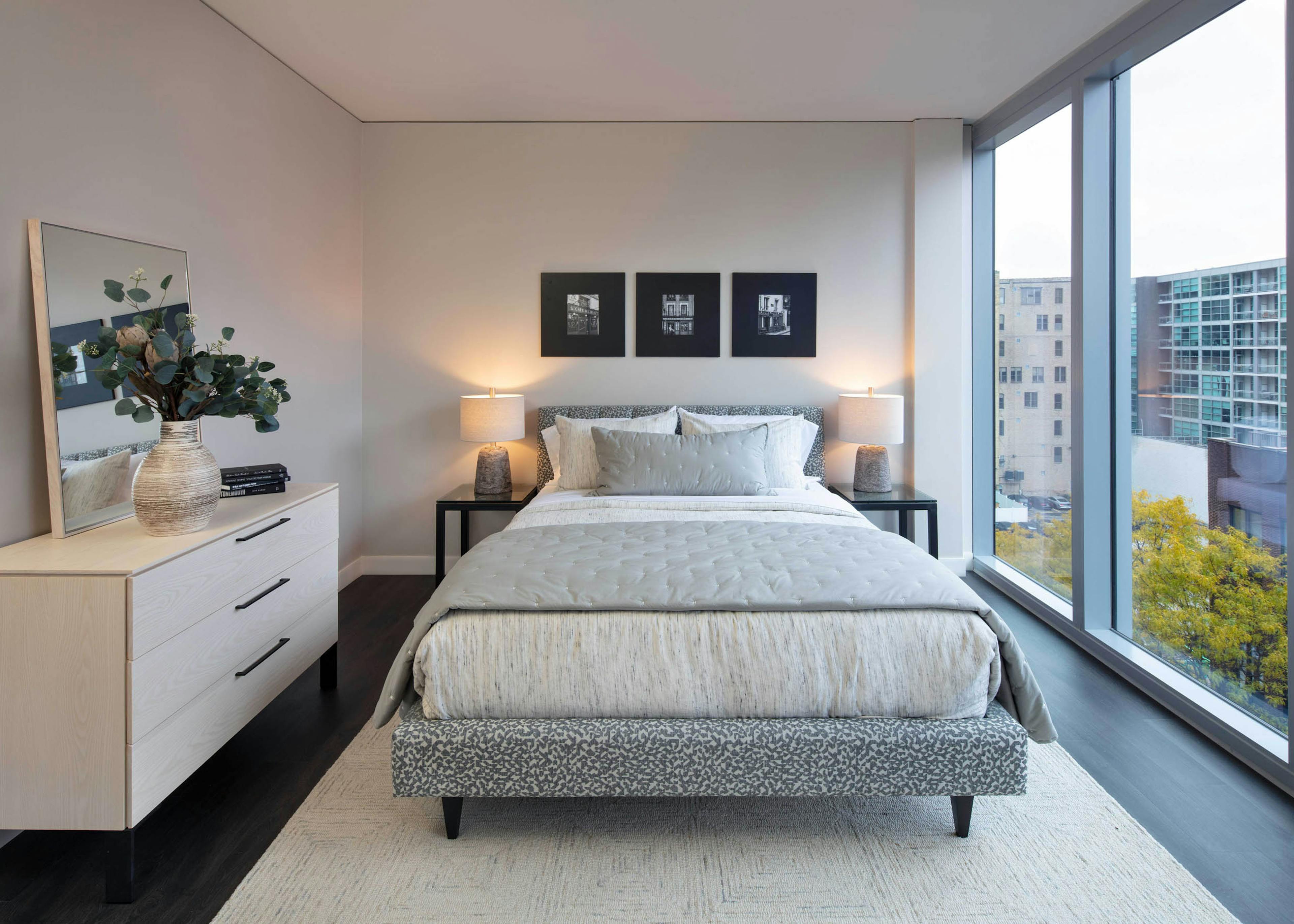 Interior shot of AMLI West Loop bedroom with a queen-sized bed, white dresser with mirror, dark wood flooring, and large window with a city view.