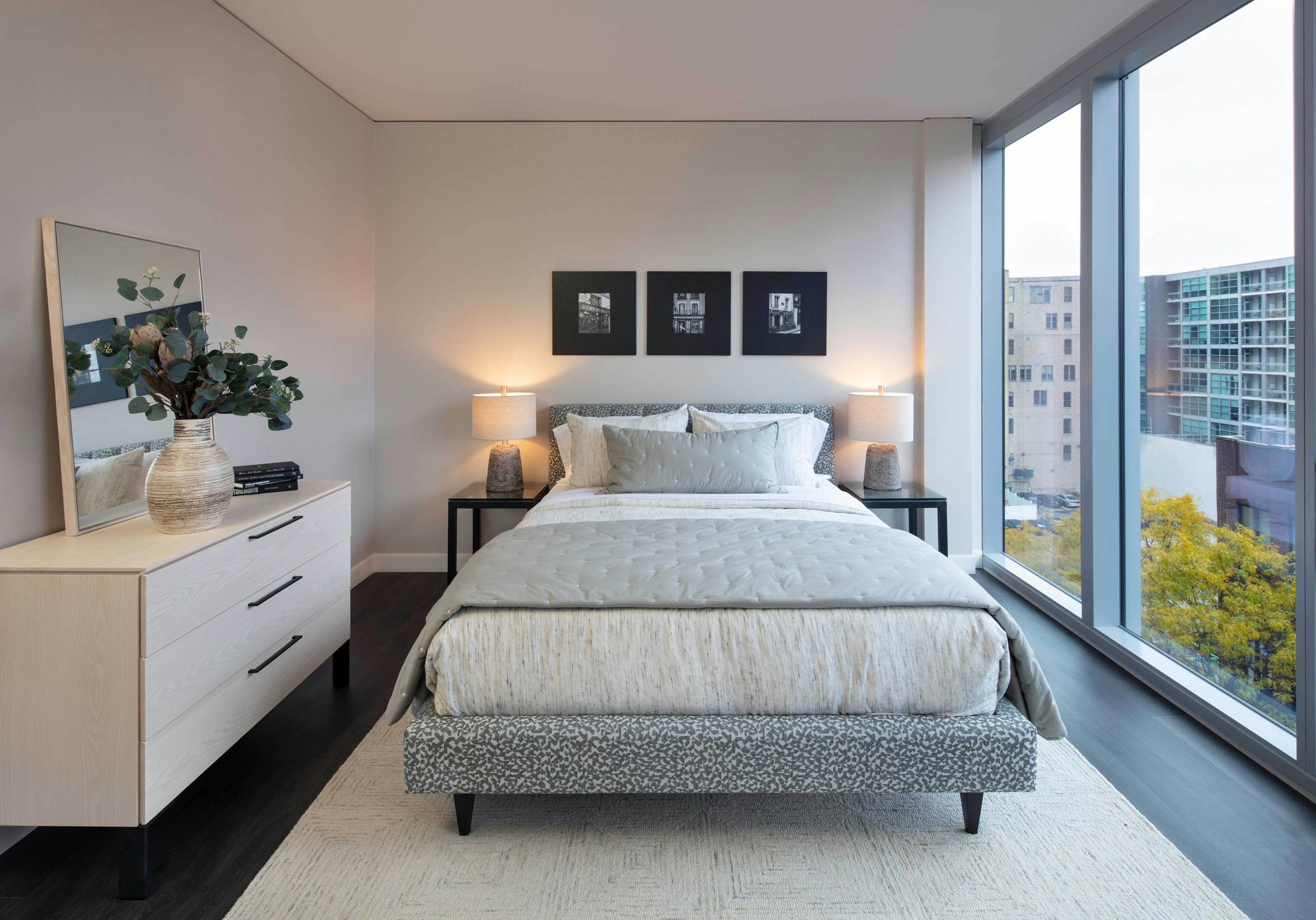 Interior shot of AMLI West Loop bedroom with a queen-sized bed, white dresser with mirror, dark wood flooring, and large window with a city view.