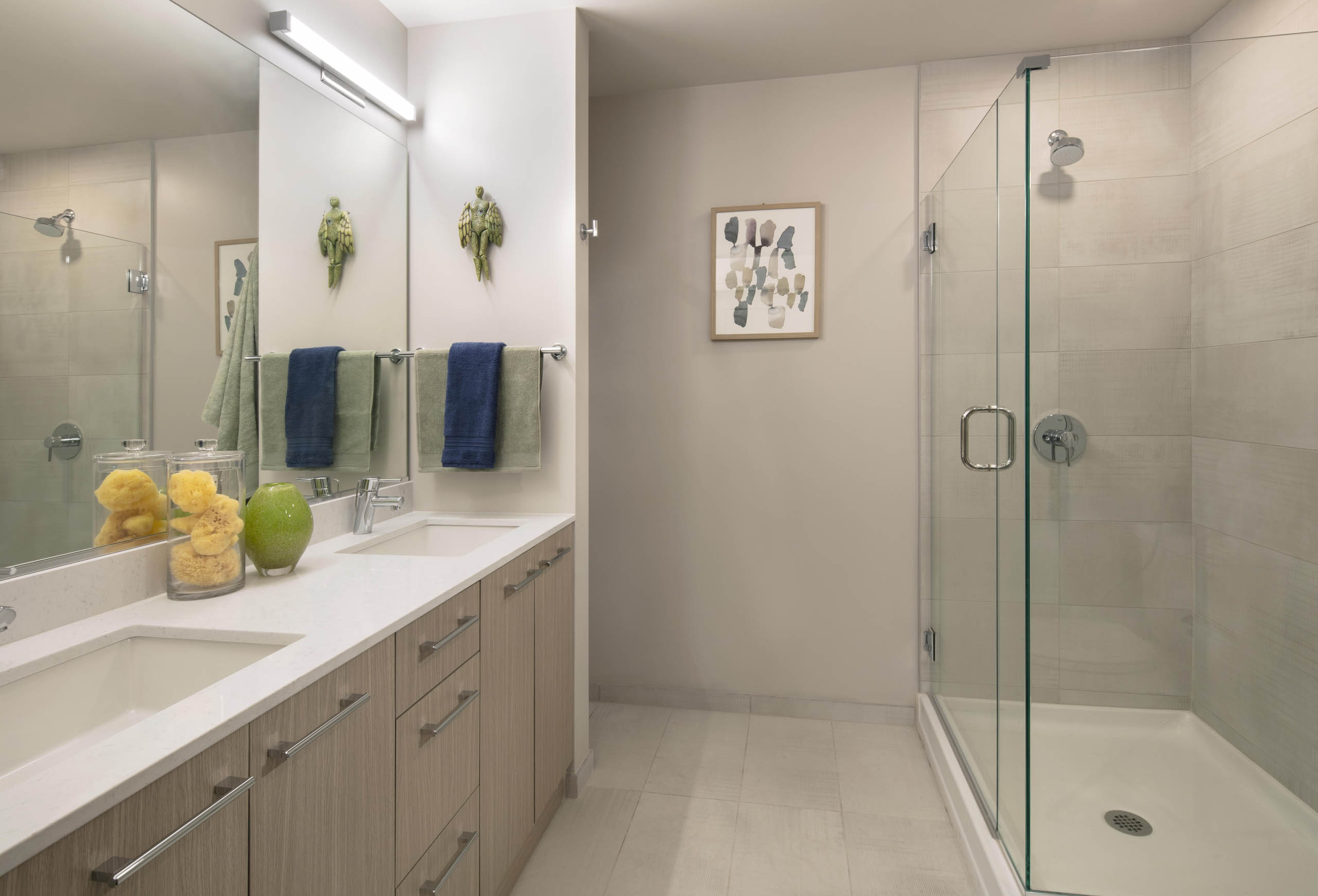 AMLI West Loop modern bathroom with a double vanity, light wood cabinets, and a walk-in shower with a glass door and tiled walls.