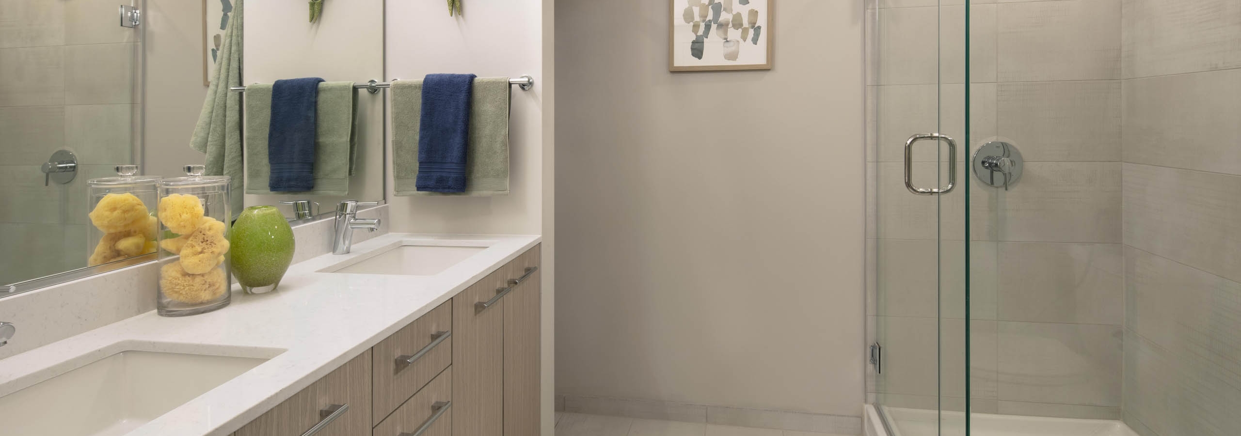 AMLI West Loop modern bathroom with a double vanity, light wood cabinets, and a walk-in shower with a glass door and tiled walls.