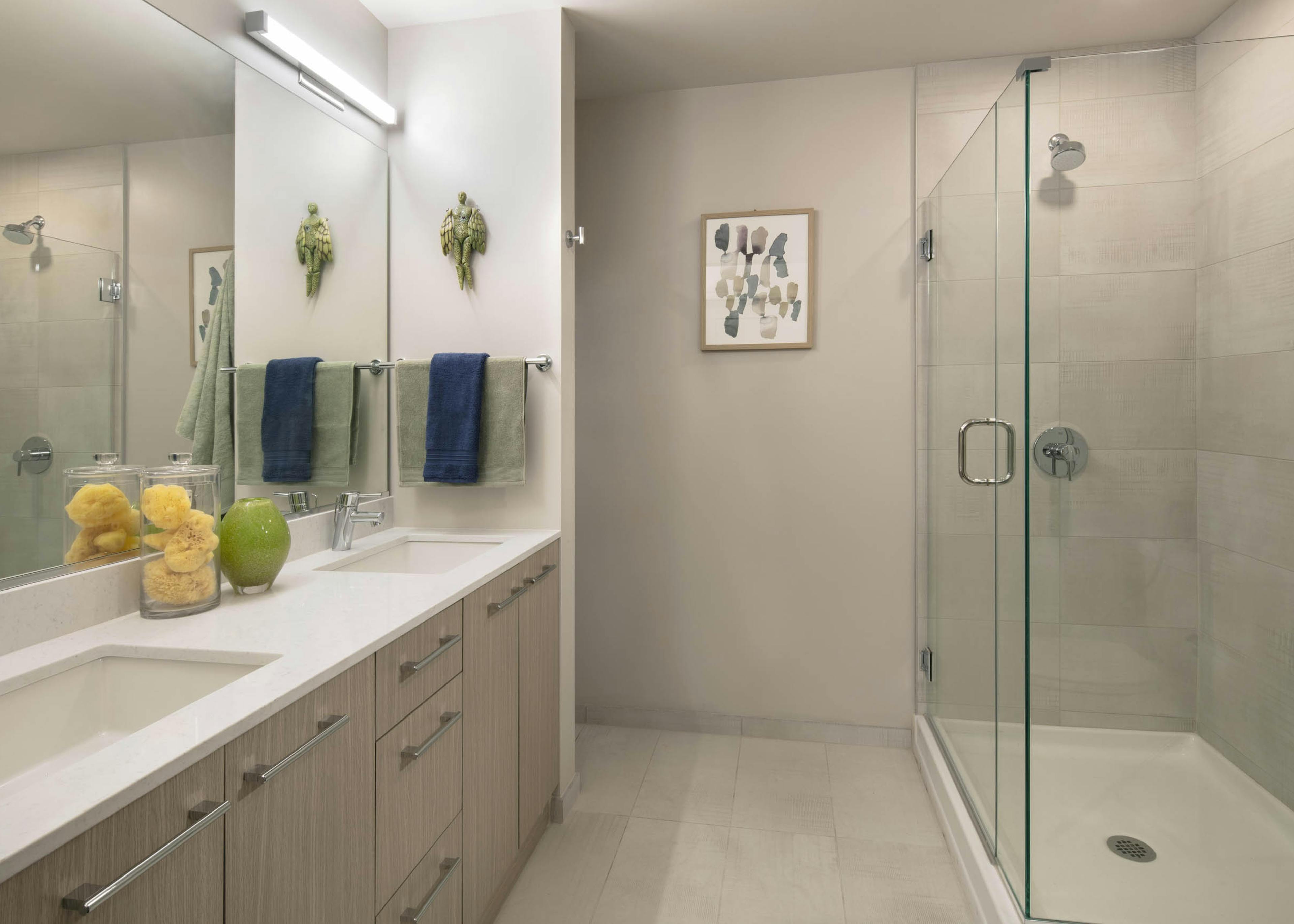 AMLI West Loop modern bathroom with a double vanity, light wood cabinets, and a walk-in shower with a glass door and tiled walls.