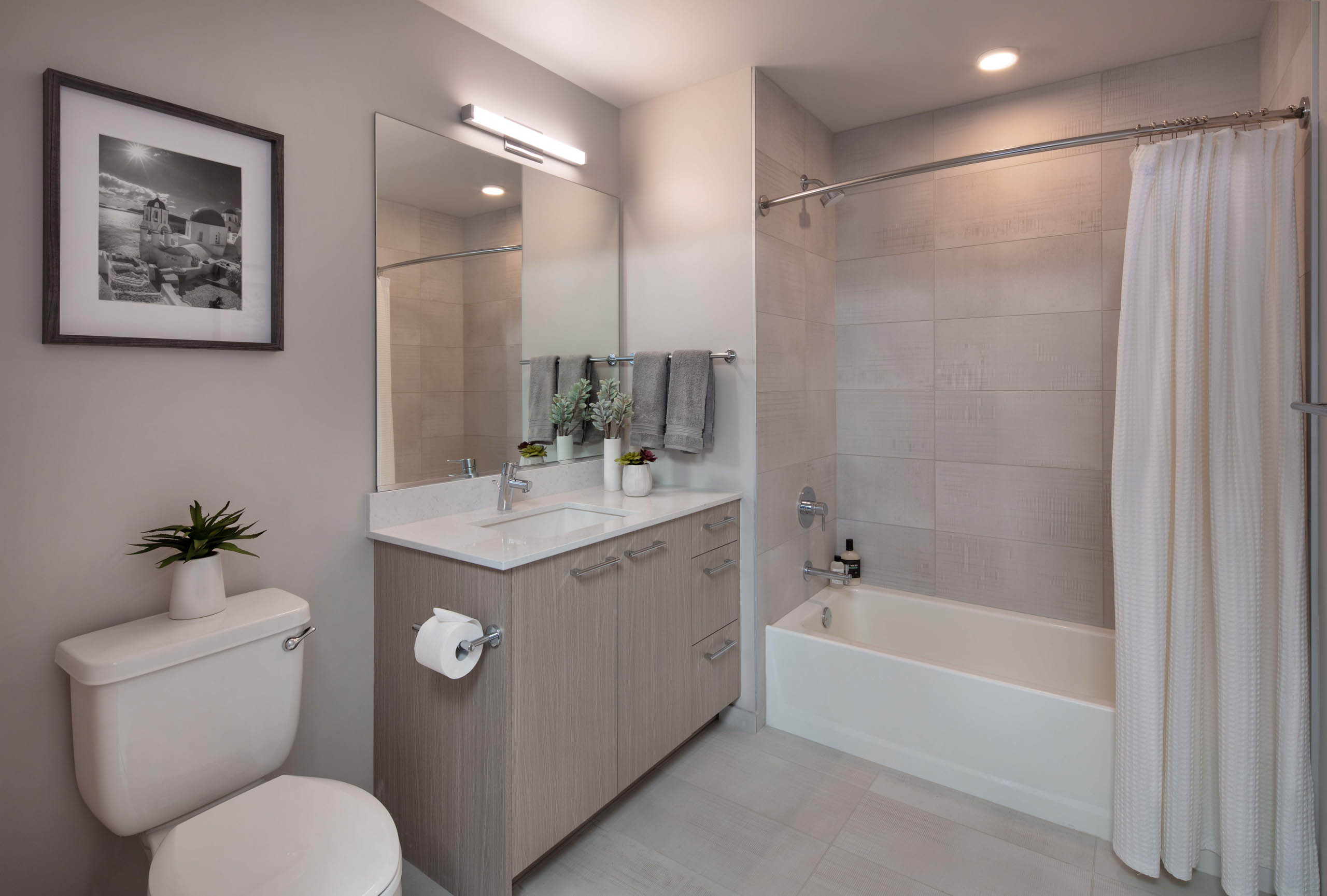 Bathroom at AMLI West Loop with black and white artwork and light wood cabinet and a white bathtub with a shower curtain and oversized tile
