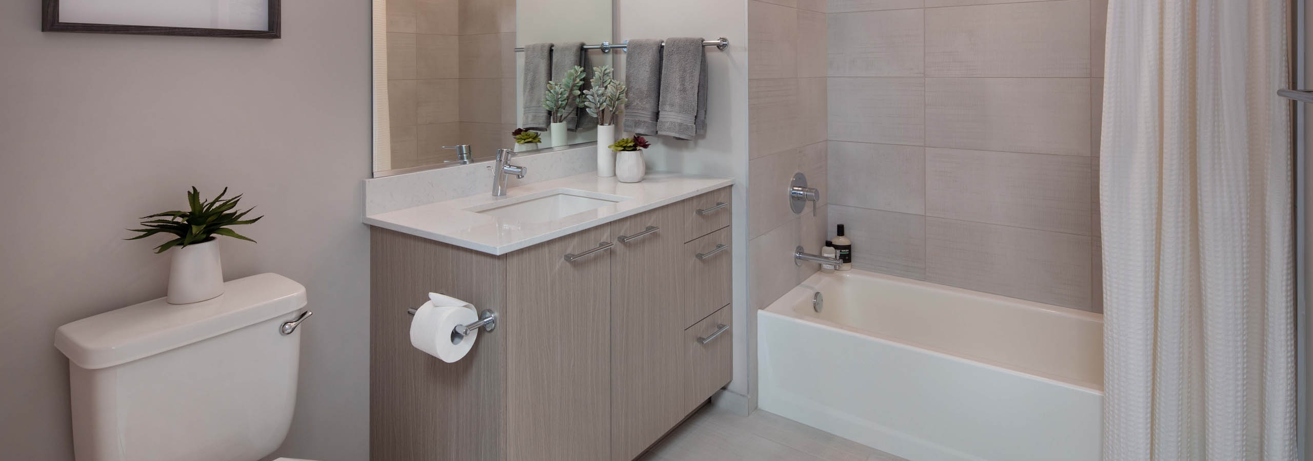 Bathroom at AMLI West Loop with black and white artwork and light wood cabinet and a white bathtub with a shower curtain and oversized tile
