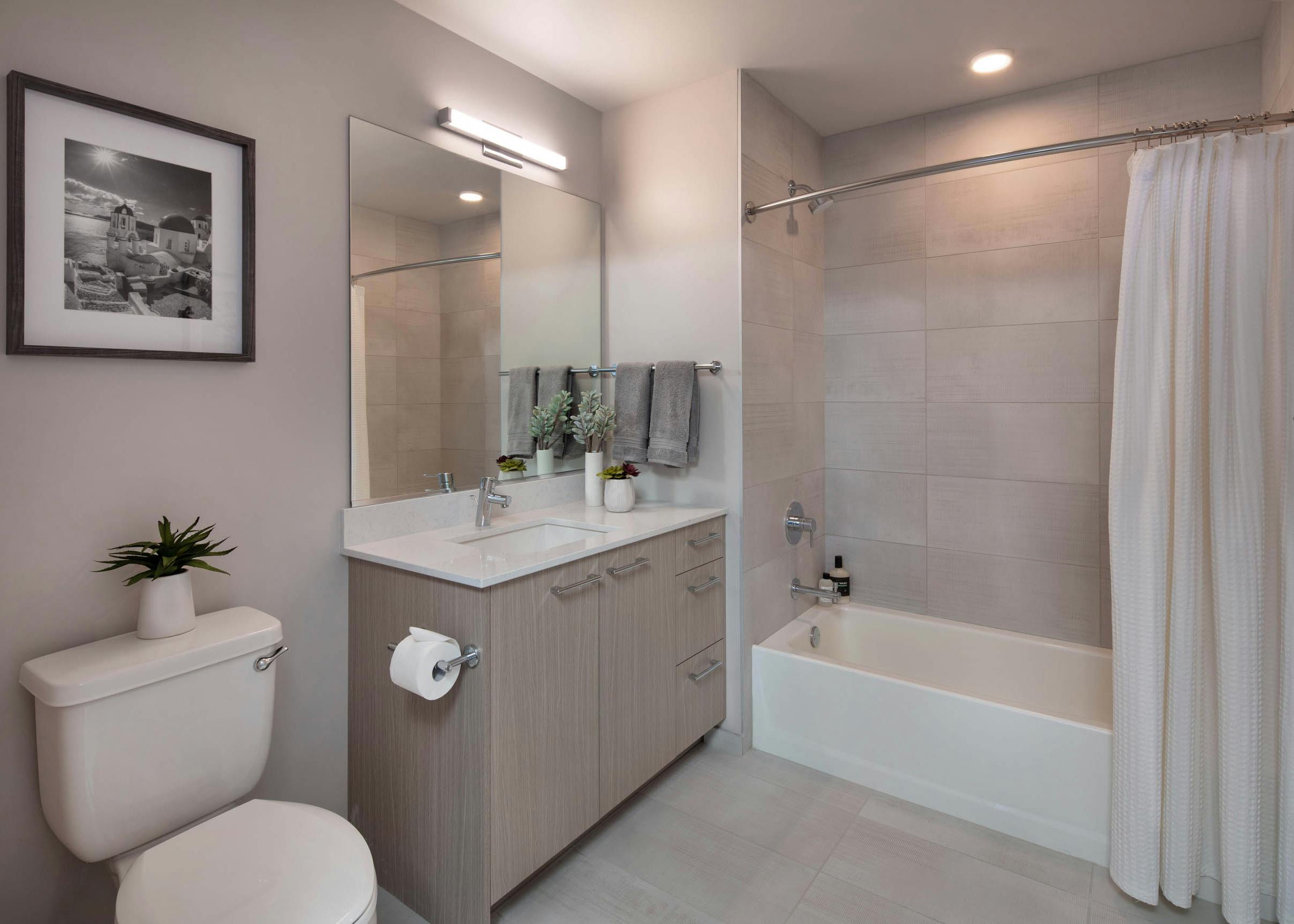 Bathroom at AMLI West Loop with black and white artwork and light wood cabinet and a white bathtub with a shower curtain and oversized tile