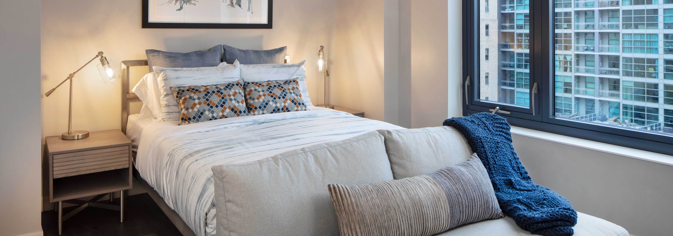 Interior view of AMLI West Loop studio living area featuring a bed, a small couch in the foreground, nightstands, and a large window with a city view.