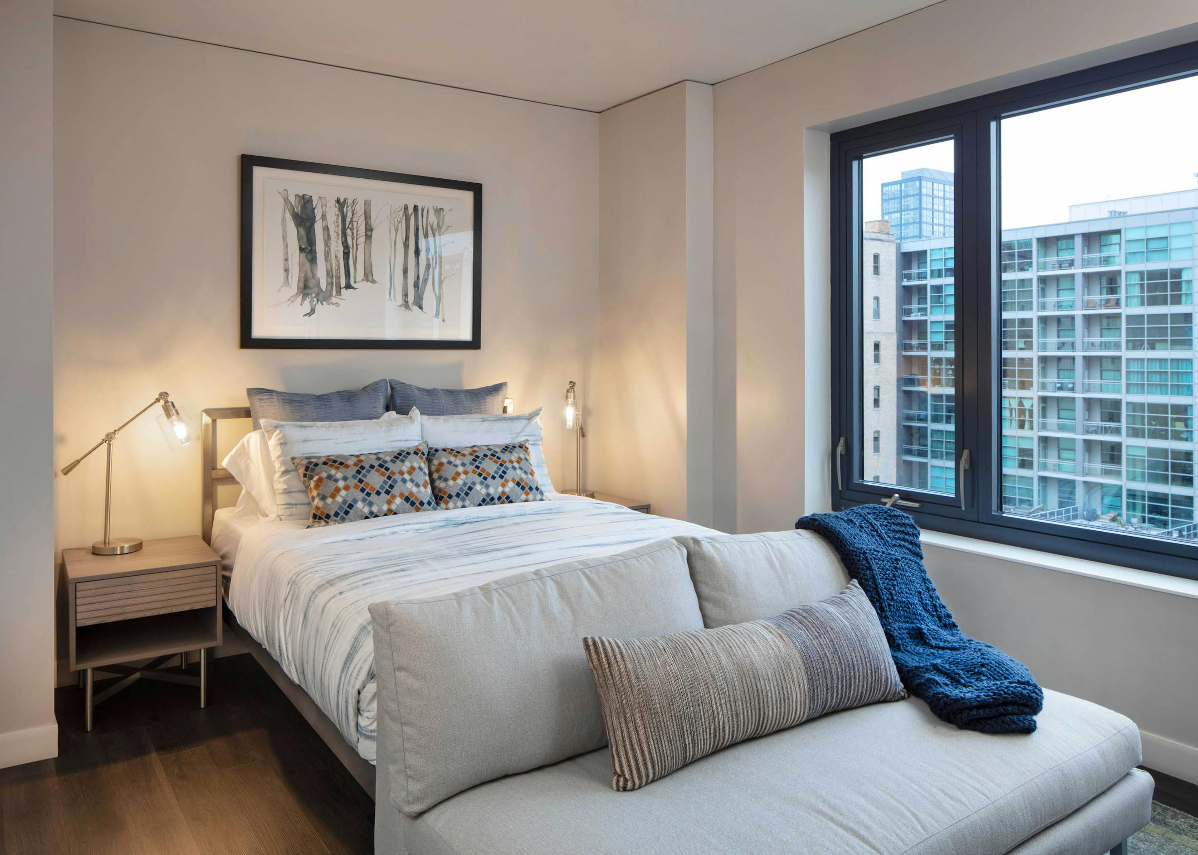 Interior view of AMLI West Loop studio living area featuring a bed, a small couch in the foreground, nightstands, and a large window with a city view.