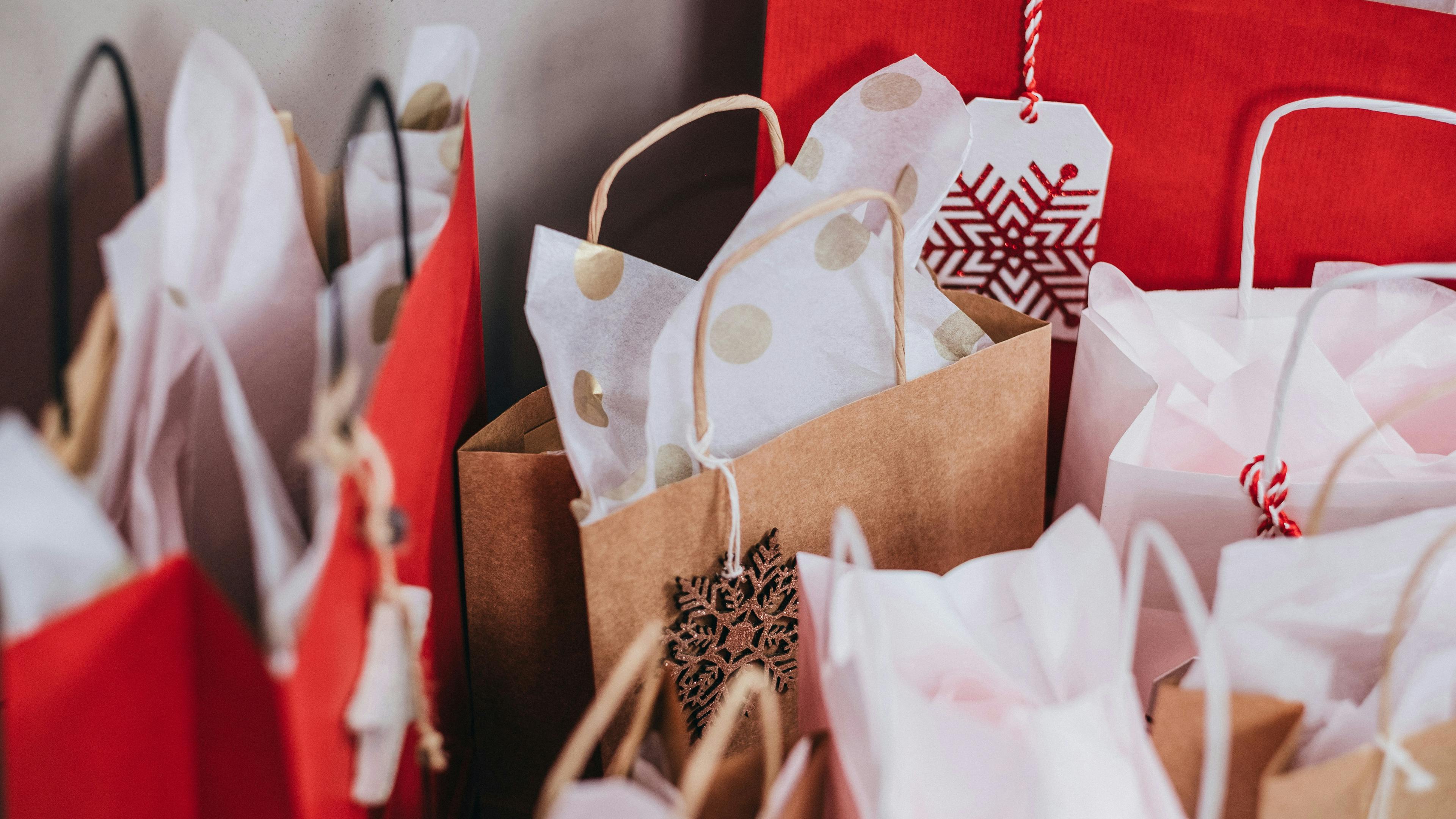 A close-up collection of brown, red, and white gift bags with tissue paper peaking out, suggesting holiday or special occasion shopping. A small wooden snowflake decoration hangs from one of the tags.
