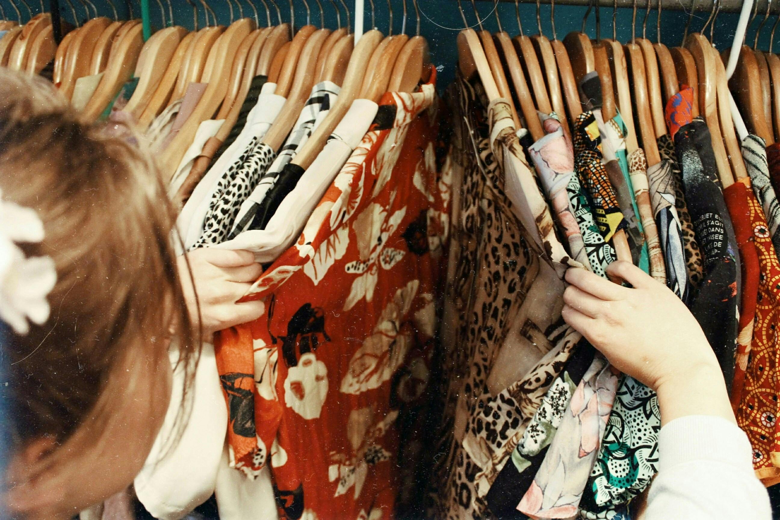 Close-up of two people's hands sorting through a rack of clothes on wooden hangers, searching for a garment. The clothes are mostly patterned, including leopard print and floral designs.