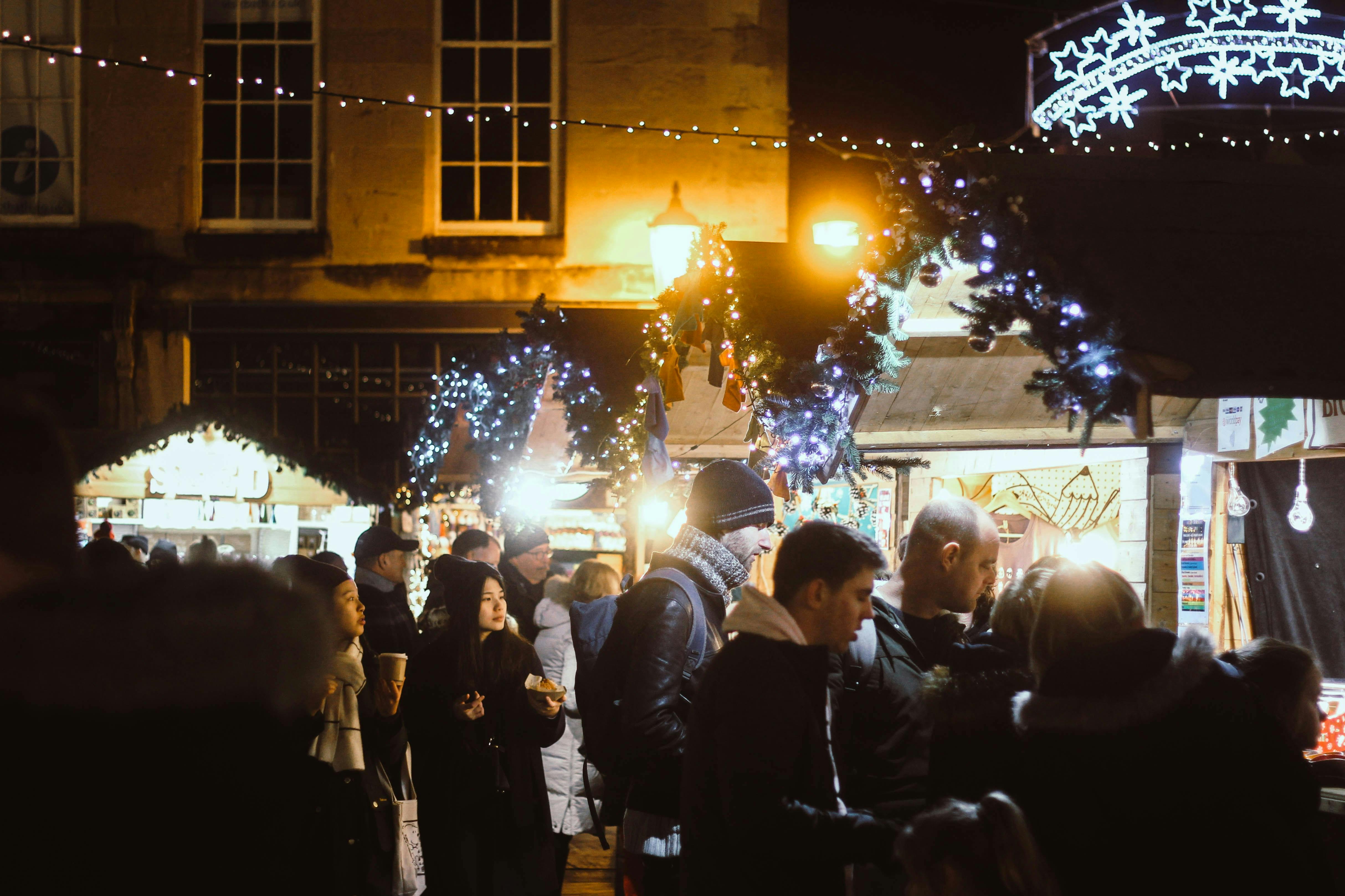 A busy, nighttime outdoor holiday or Christmas market with many people gathered. The stalls and buildings are decorated with string lights, wreaths, and festive white lights.