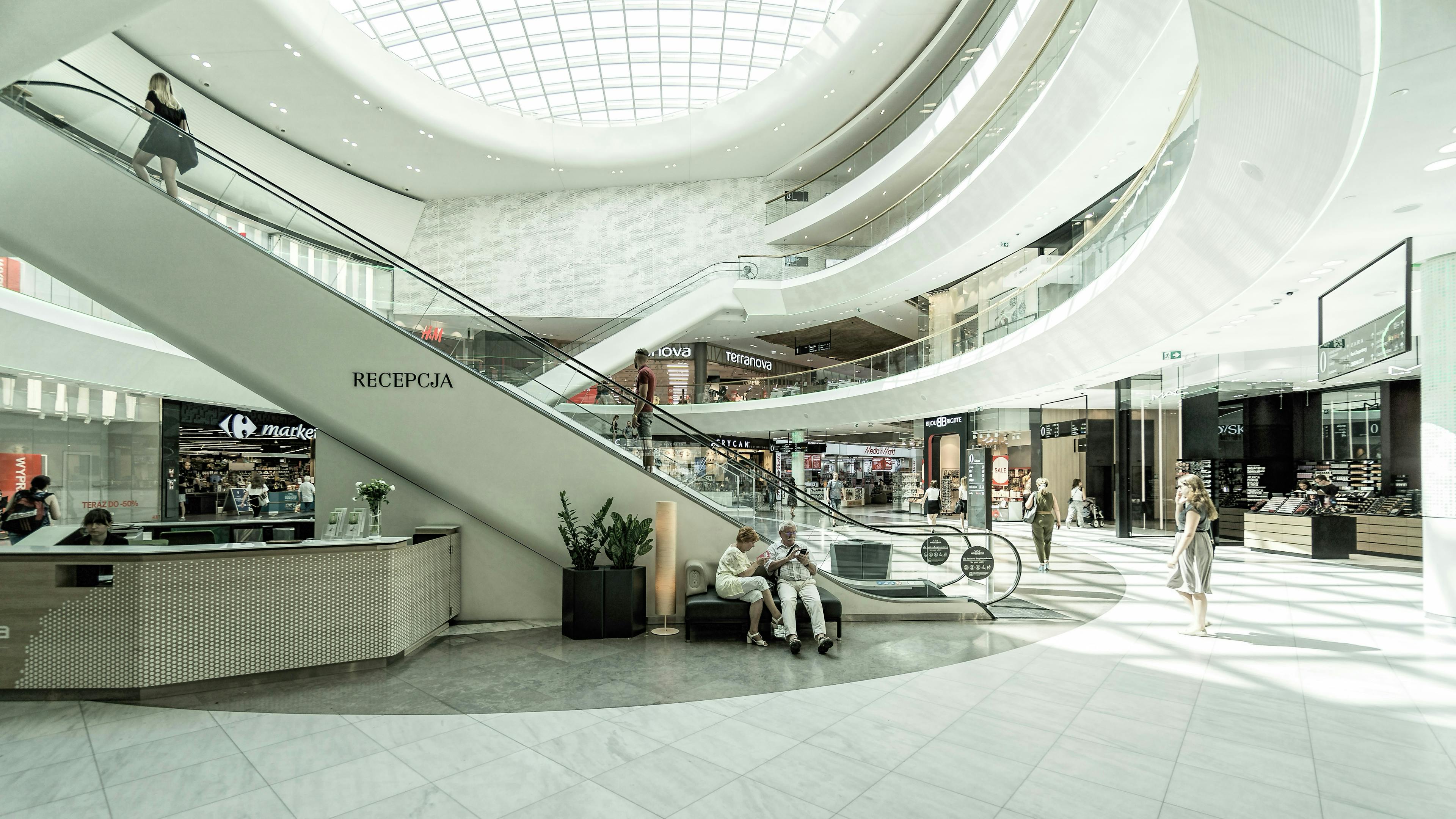 The bright, modern, and spacious white atrium of a shopping mall, featuring a large, curved escalator leading up. Shoppers are scattered throughout, and a group is seated near a reception desk at the base of the escalator.
