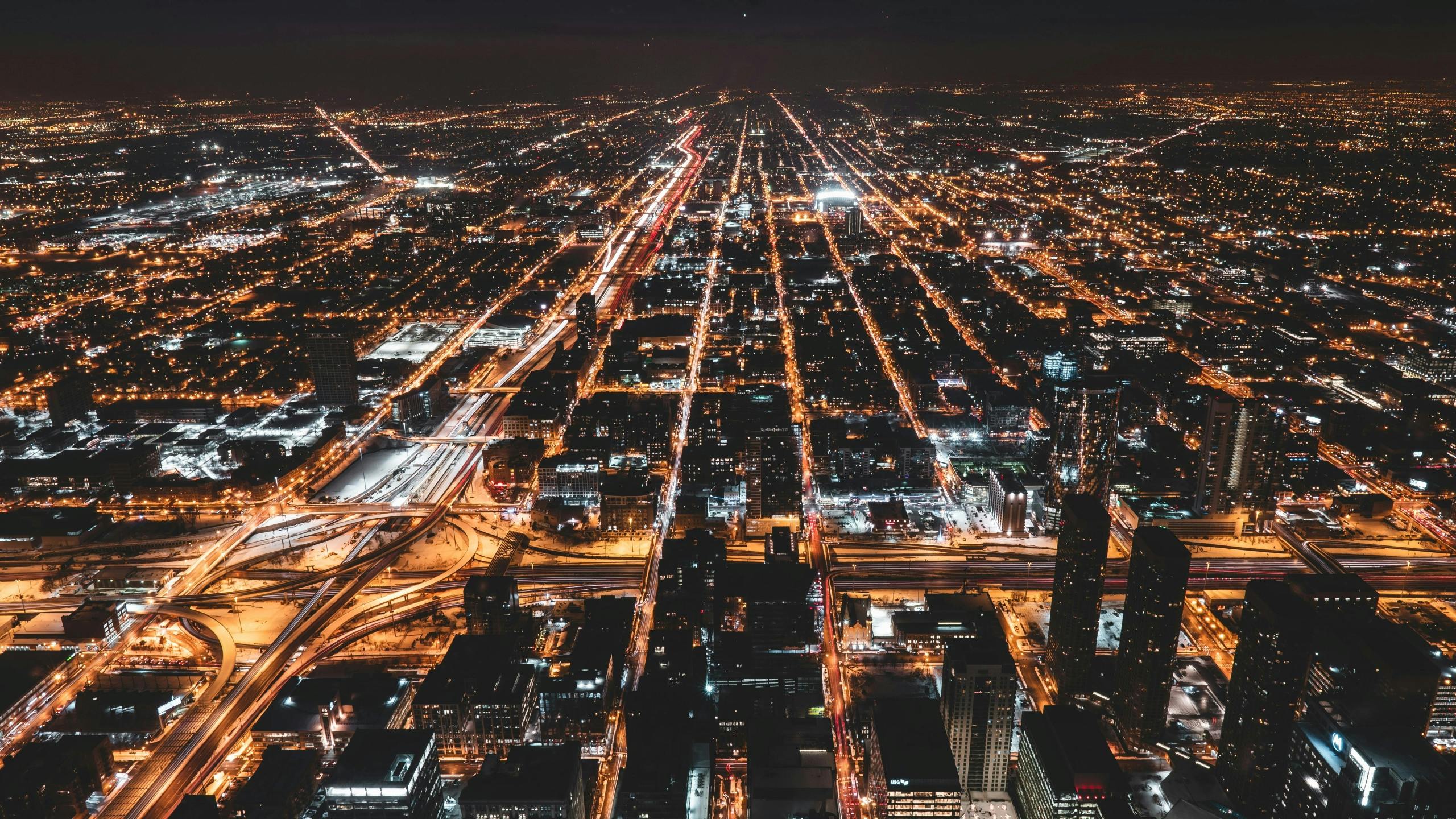 High-angle nighttime aerial view of the West Loop neighborhood of Chicago, showing bright street grids and major expressway interchanges.