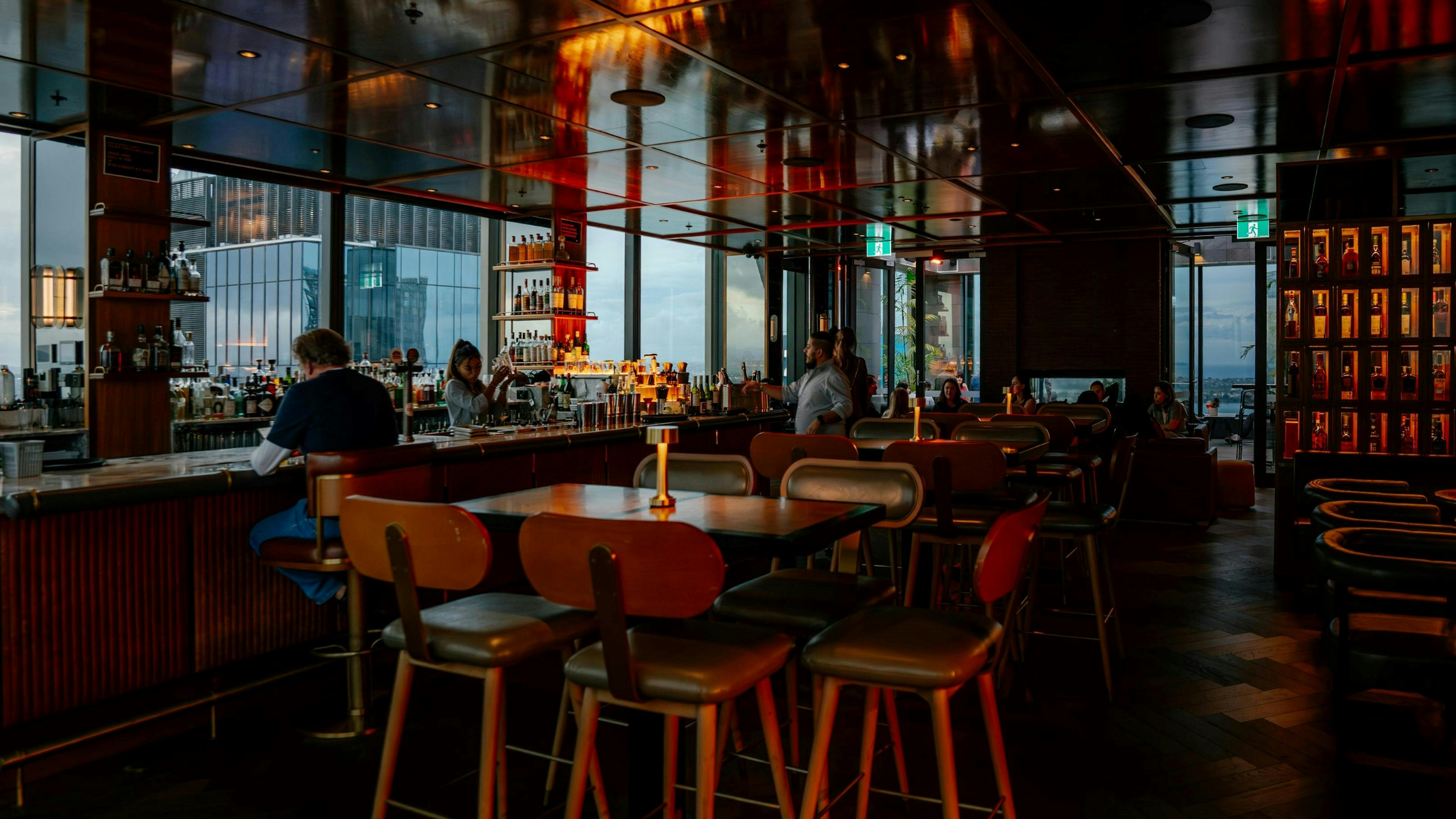 Interior of a dimly lit, upscale bar with a reflective ceiling, featuring high-top tables, leather seating, and a busy bar area next to large windows overlooking cityscape.
