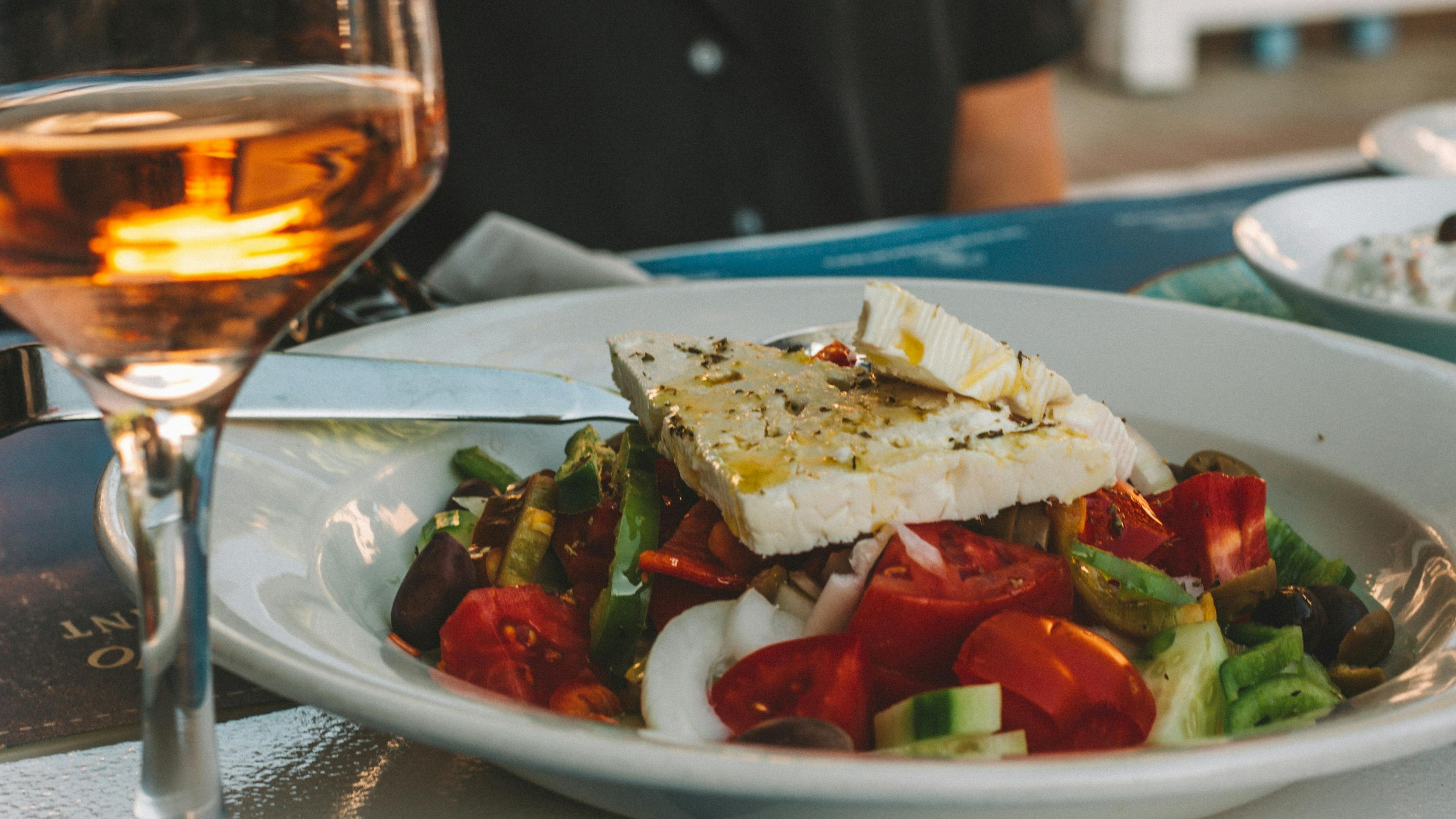 A close-up of a white plate with Greek salad topped with a large slice of feta cheese, next to a glass of wine.