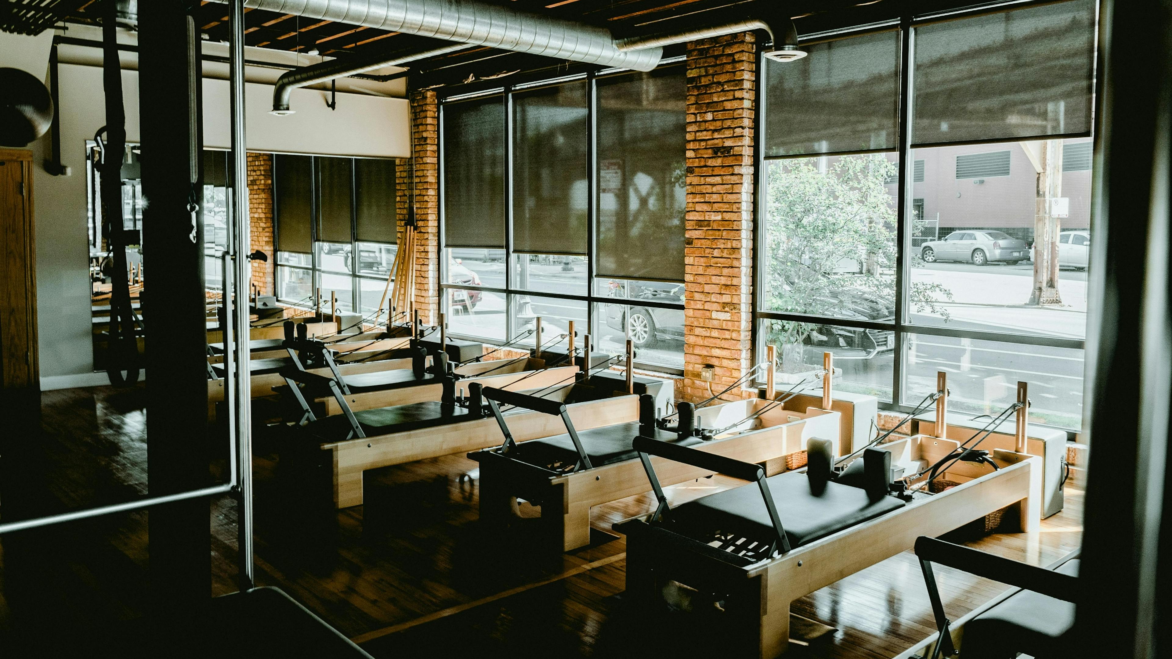 Interior of a Pilates studio with a row of wooden Reformer machines, exposed brick, and large windows with street views.