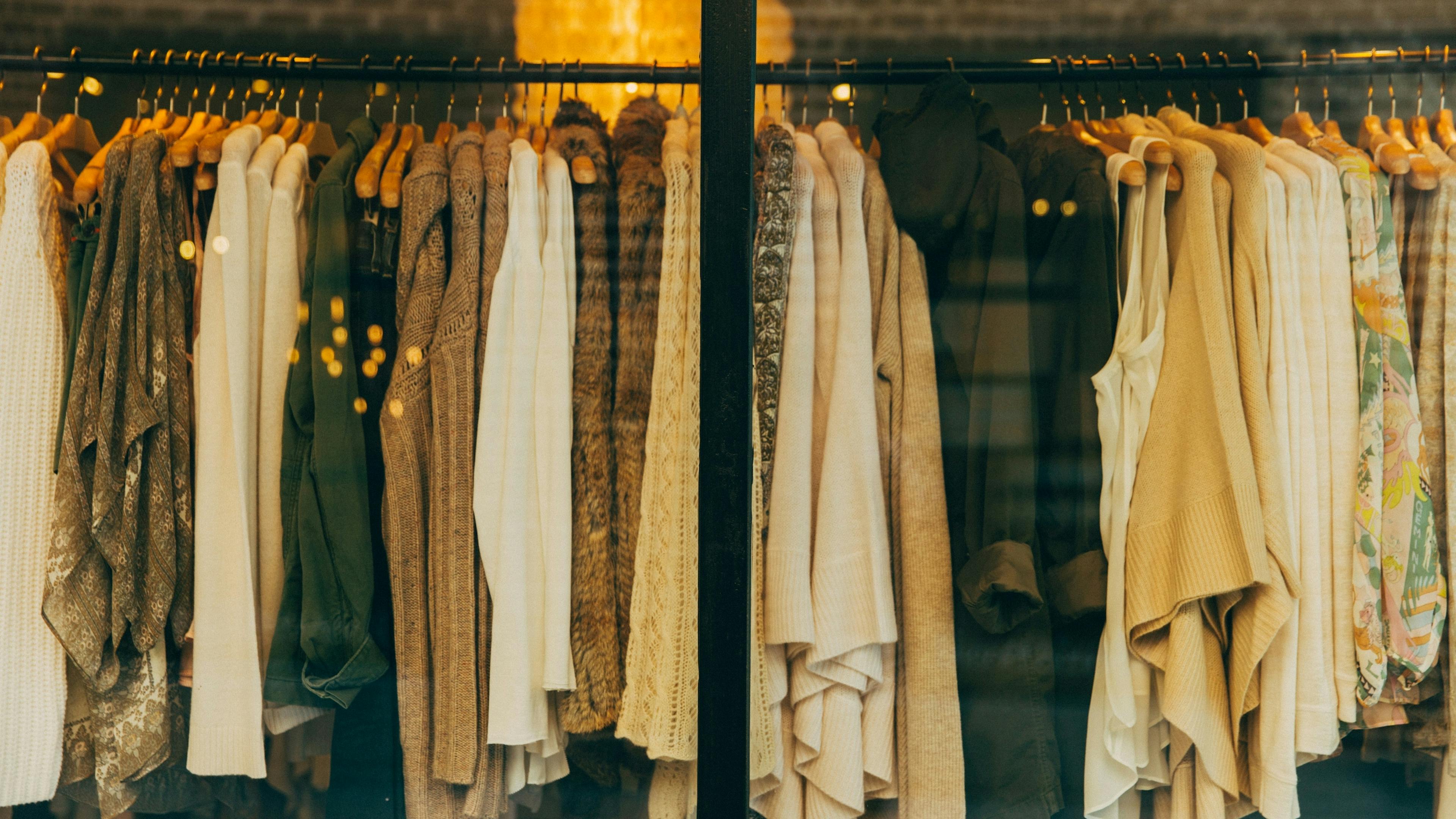 Close-up of a clothing rack filled with garments in natural, earth-tone colors like white, beige, brown, and green, seen through a store window.