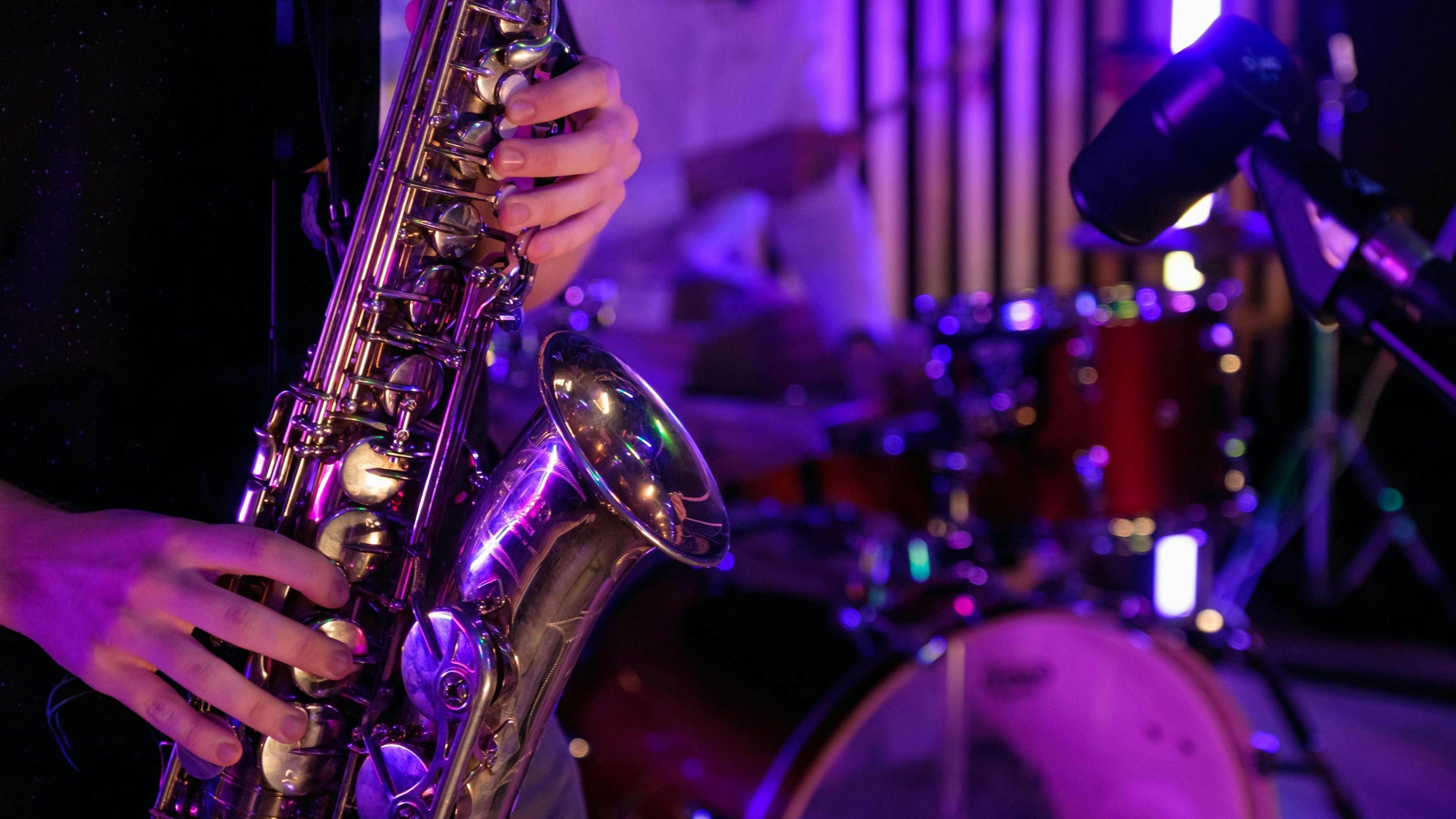 Close-up of a person playing a saxophone under purple stage lighting with a drum set and another musician blurred in the background.