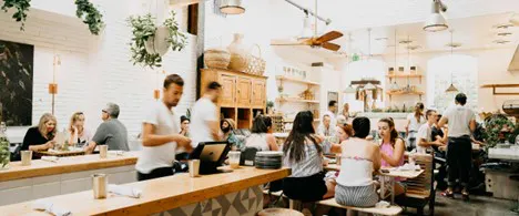 interior photo of a bright, modern, and busy restaurant with people seated at wooden tables and long bar counter