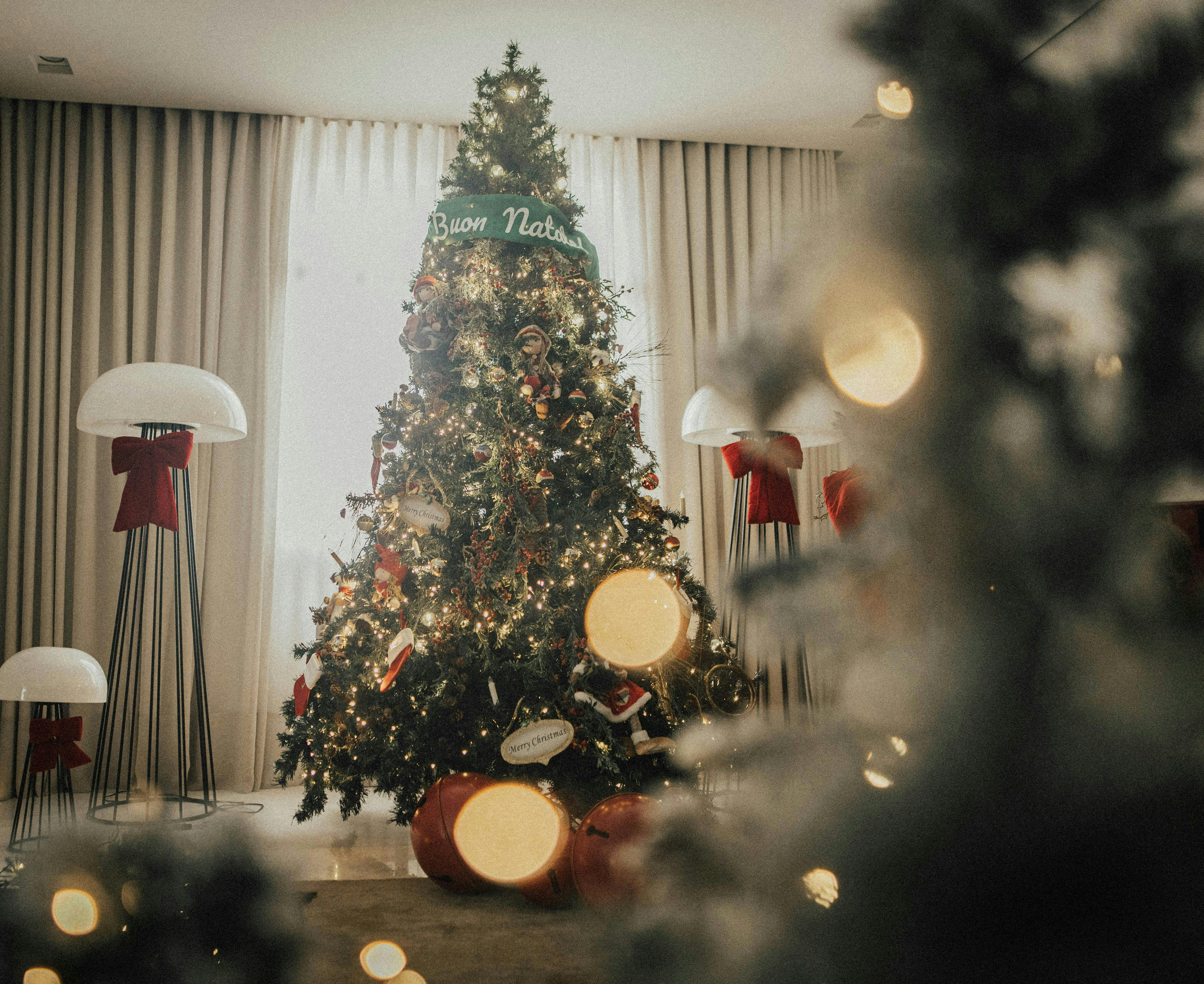 A tall, fully decorated holiday tree in a modern living room setting, featuring string lights that create bokeh effects in the foreground. The tree is decorated with a "Buon Natale" banner and stands in front of white curtains, flanked by modern floor lamps.