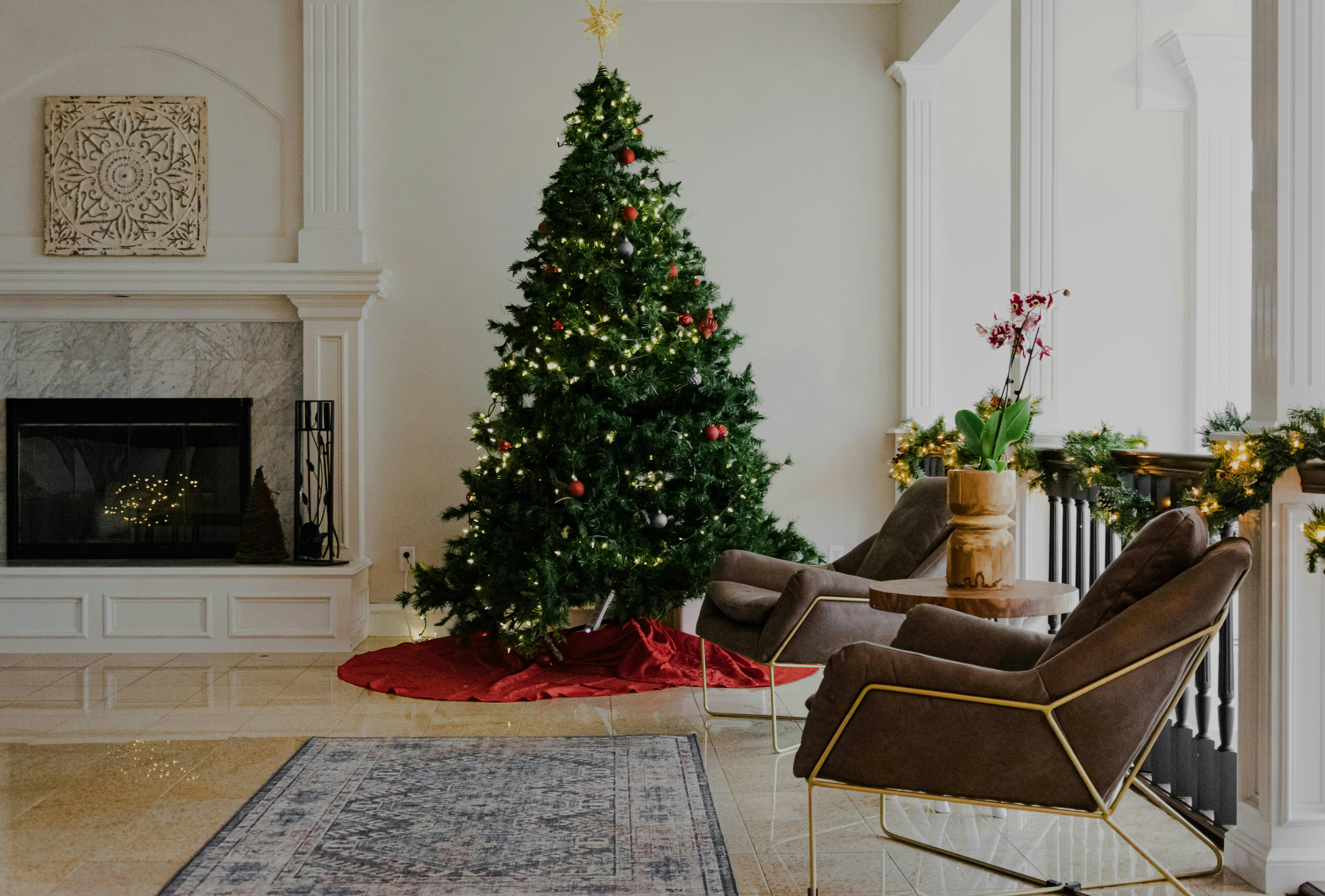 A cozy, neutral-toned living room featuring a tall, decorated holiday tree on a red tree skirt next to a marble-mantled fireplace. Two modern, low-slung armchairs sit on a patterned rug in the foreground.