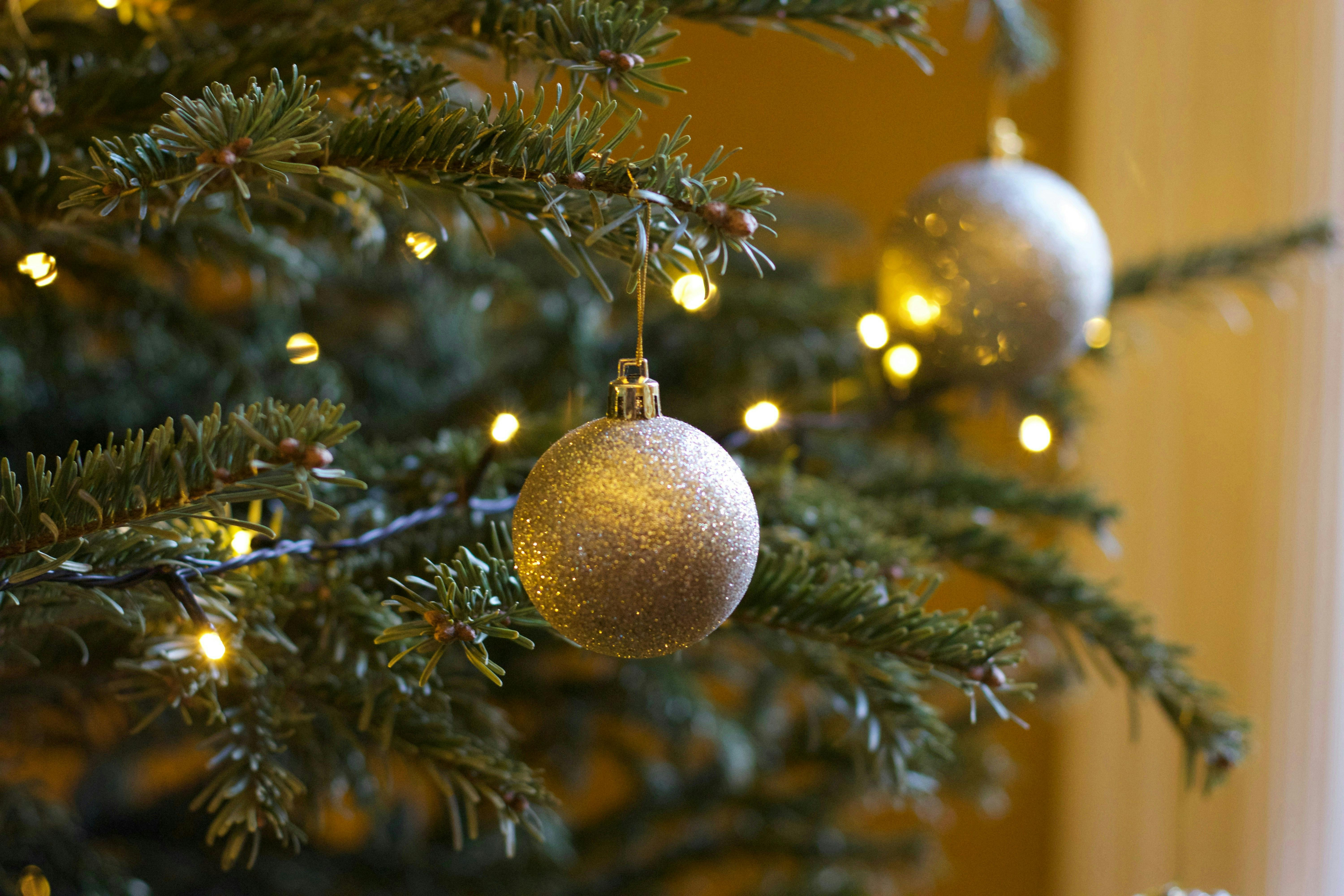 A close-up, macro shot of a holiday tree branch adorned with warm string lights. A silver, glitter-covered spherical ornament hangs prominently in the foreground.
