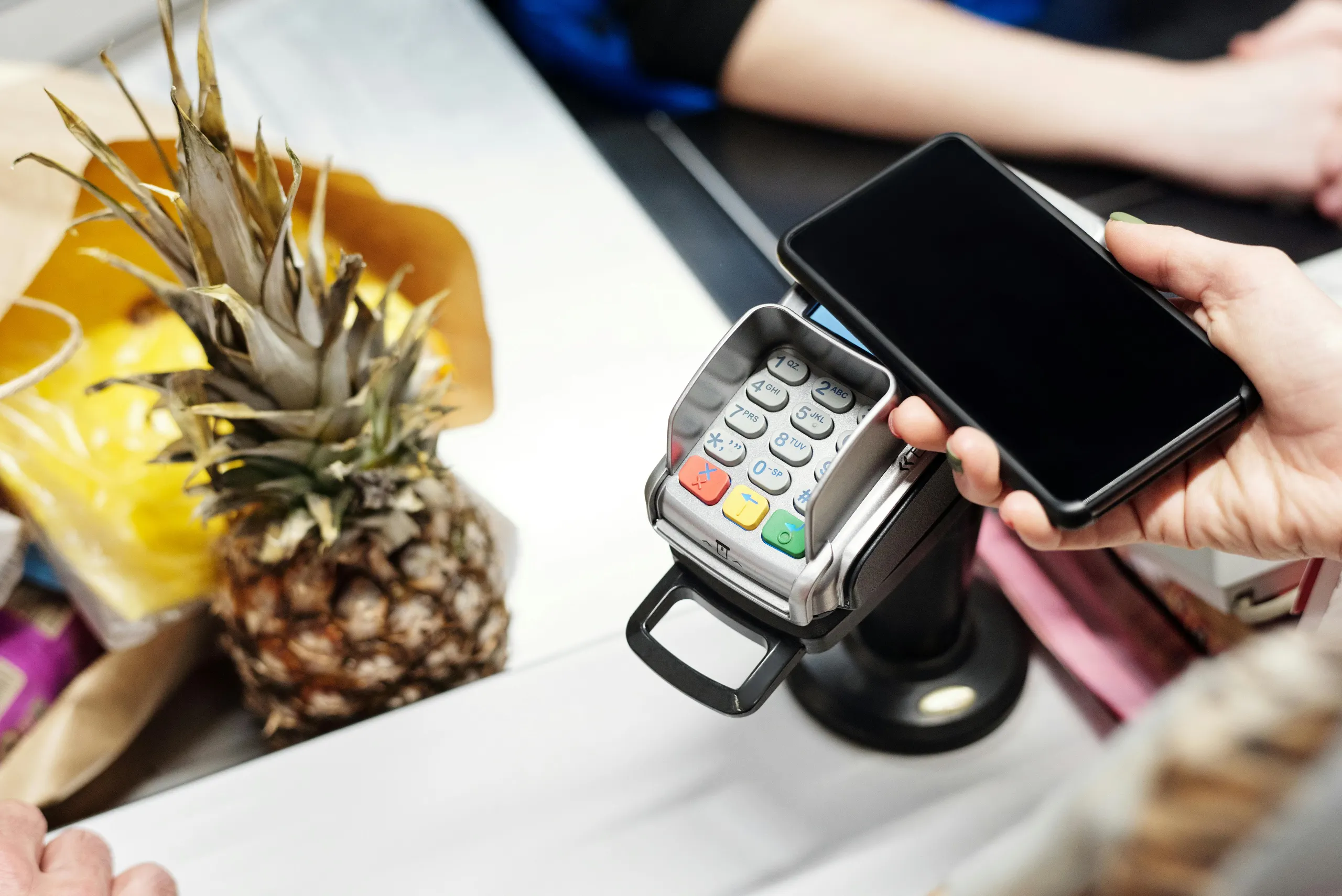 Hand holding a smartphone over a point-of-sale terminal to make a contactless payment, with a pineapple and other groceries in the background.