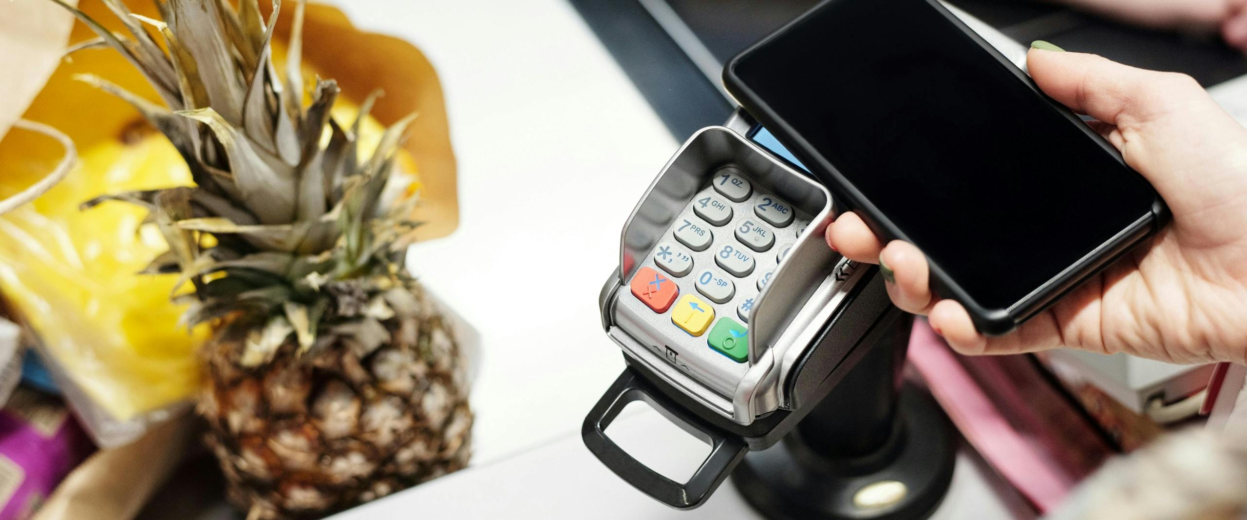 Hand holding a smartphone over a point-of-sale terminal to make a contactless payment, with a pineapple and other groceries in the background.