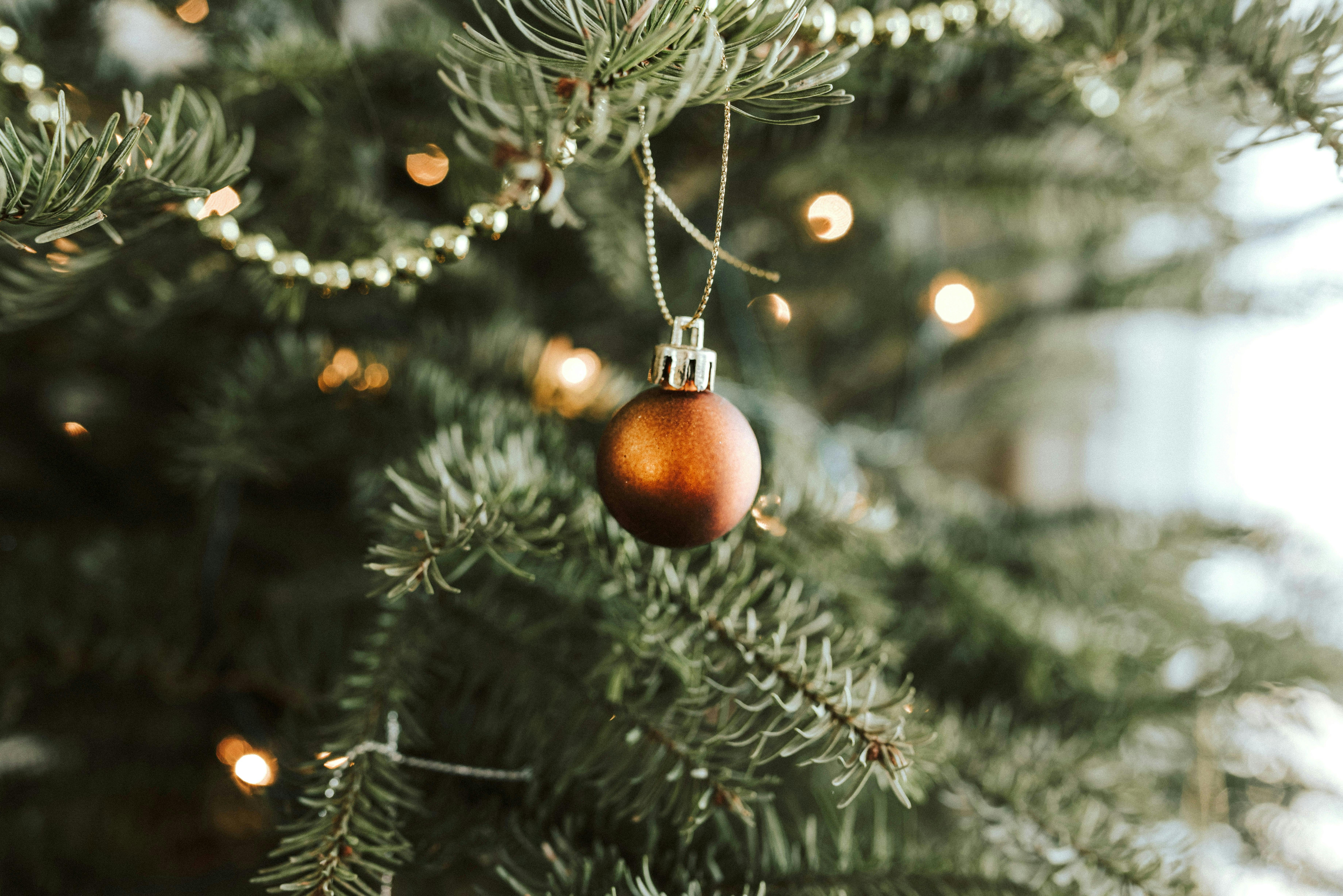 A single, matte copper-colored ball ornament hangs from a pine branch. A strand of gold beaded garland drapes above it, and the background is filled with the soft, warm bokeh of Christmas tree lights.

