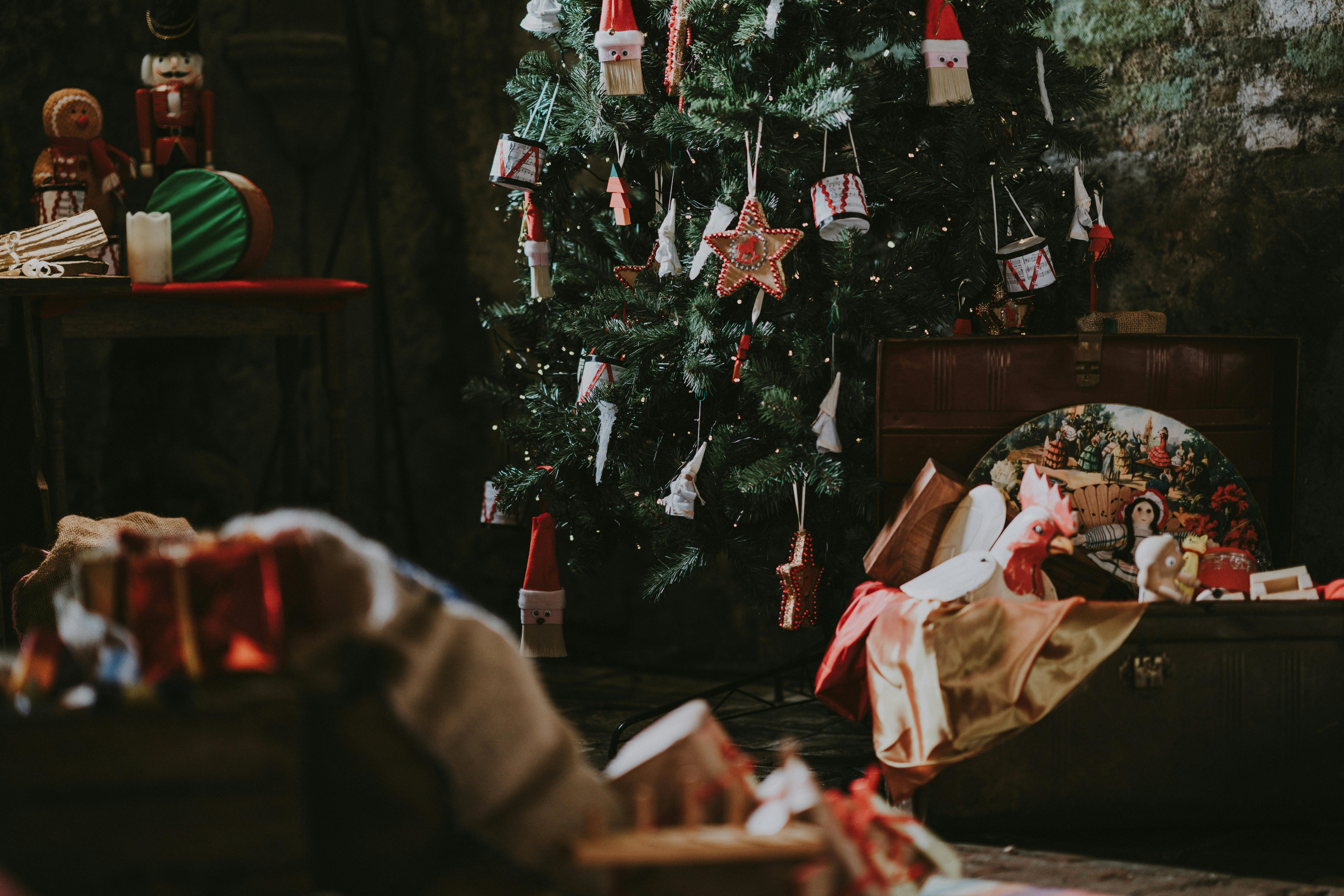 A vintage-style holiday scene featuring a tree decorated with fabric drums, stars, and Santa ornaments. To the left is a table with nutcrackers, and to the right is an open trunk filled with classic toys, including a wooden rooster.