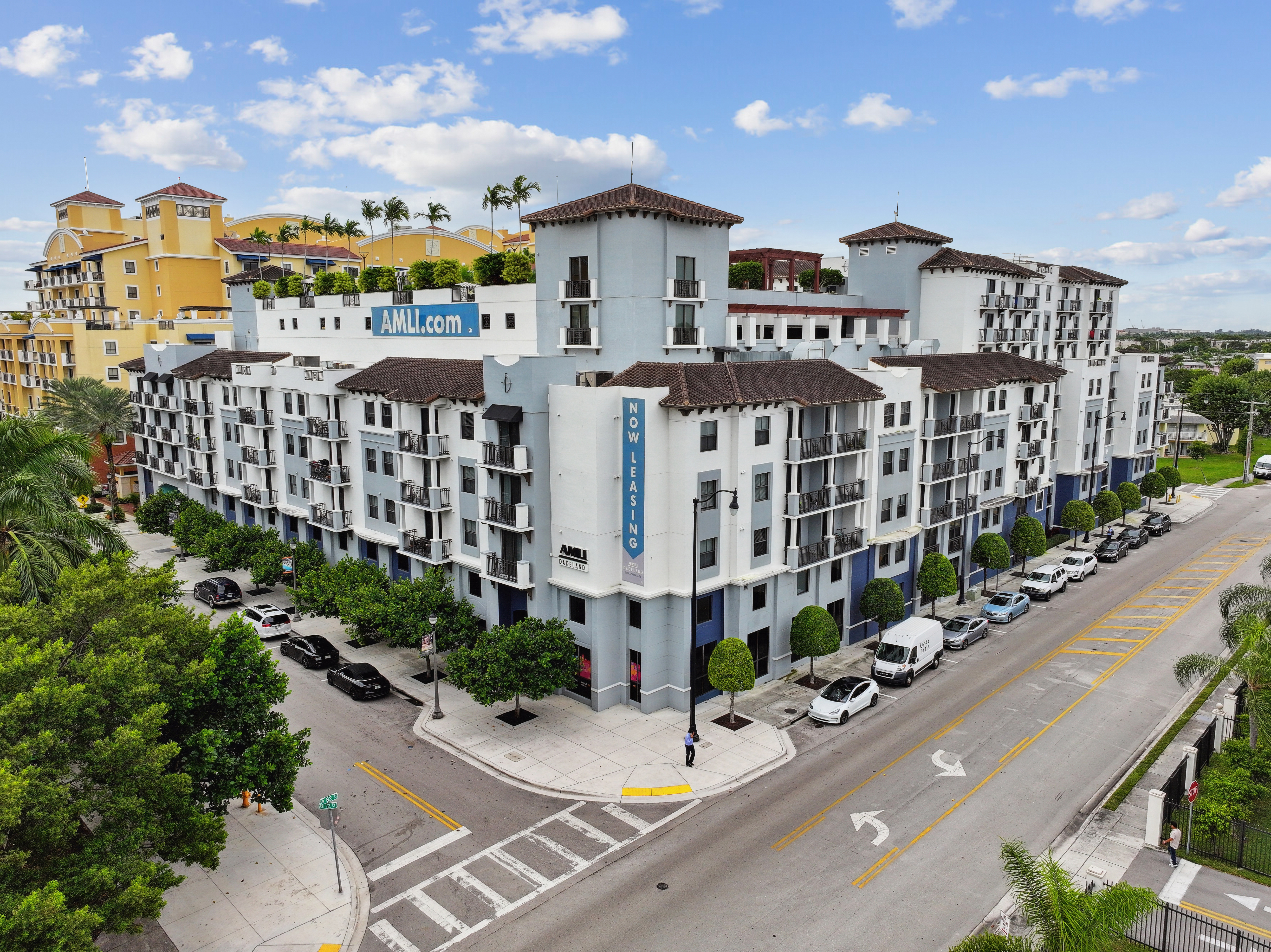 Exterior view of the AMLI Dadeland apartment building with seven floors and street parking and trees and blue sky with clouds