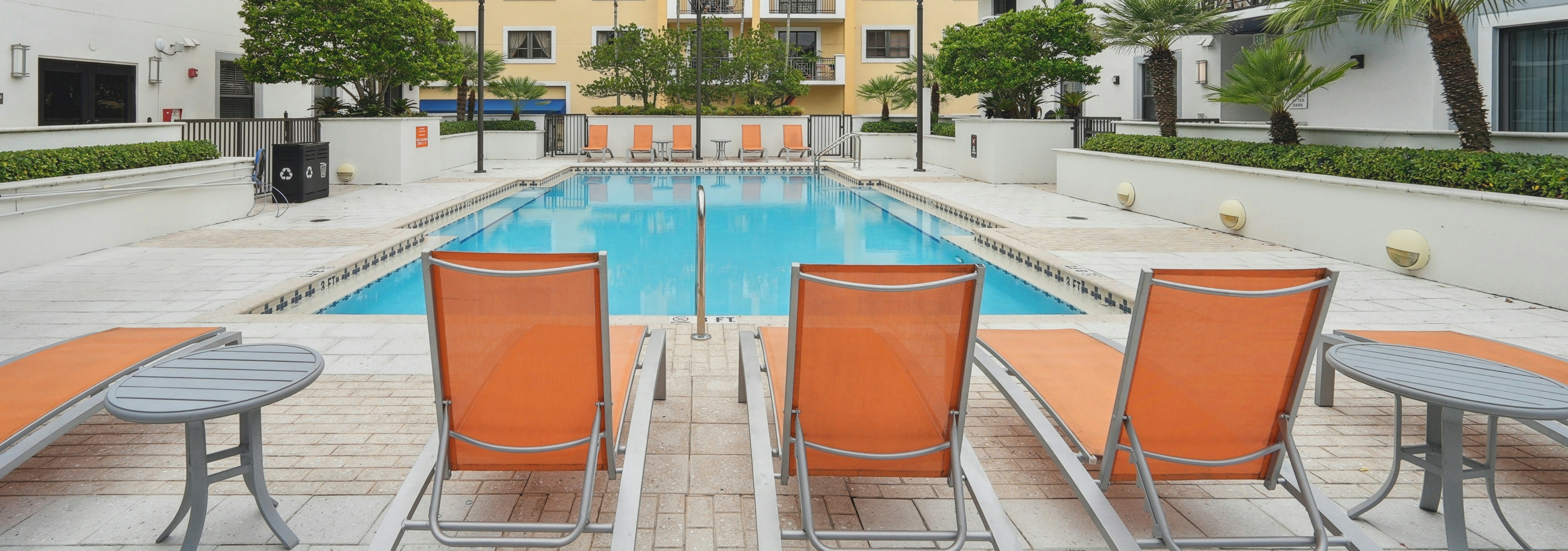 Exterior view of a swimming pool at AMLI Dadeland apartments with orange lounge chairs and lush green plants surrounding